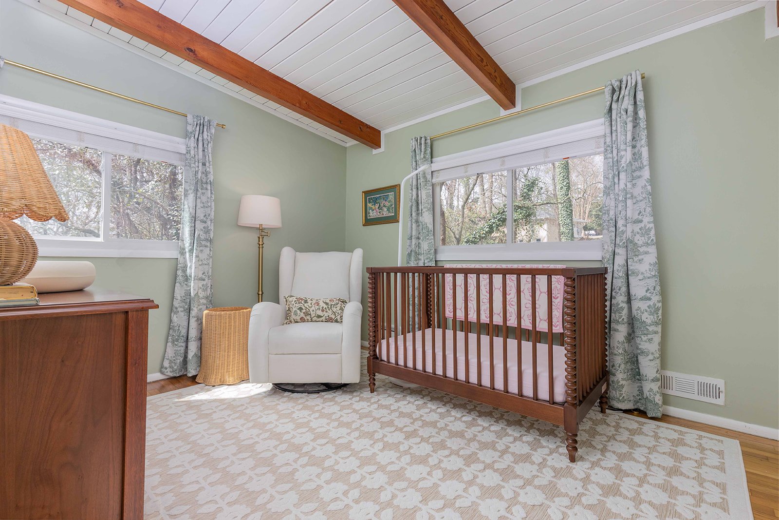 A cozy nursery with light green walls, white wooden ceiling with exposed wooden beams, and large windows with patterned curtains. There is a white armchair with a small decorative pillow, a wooden crib with a pink mattress and a pink patterned sheet,