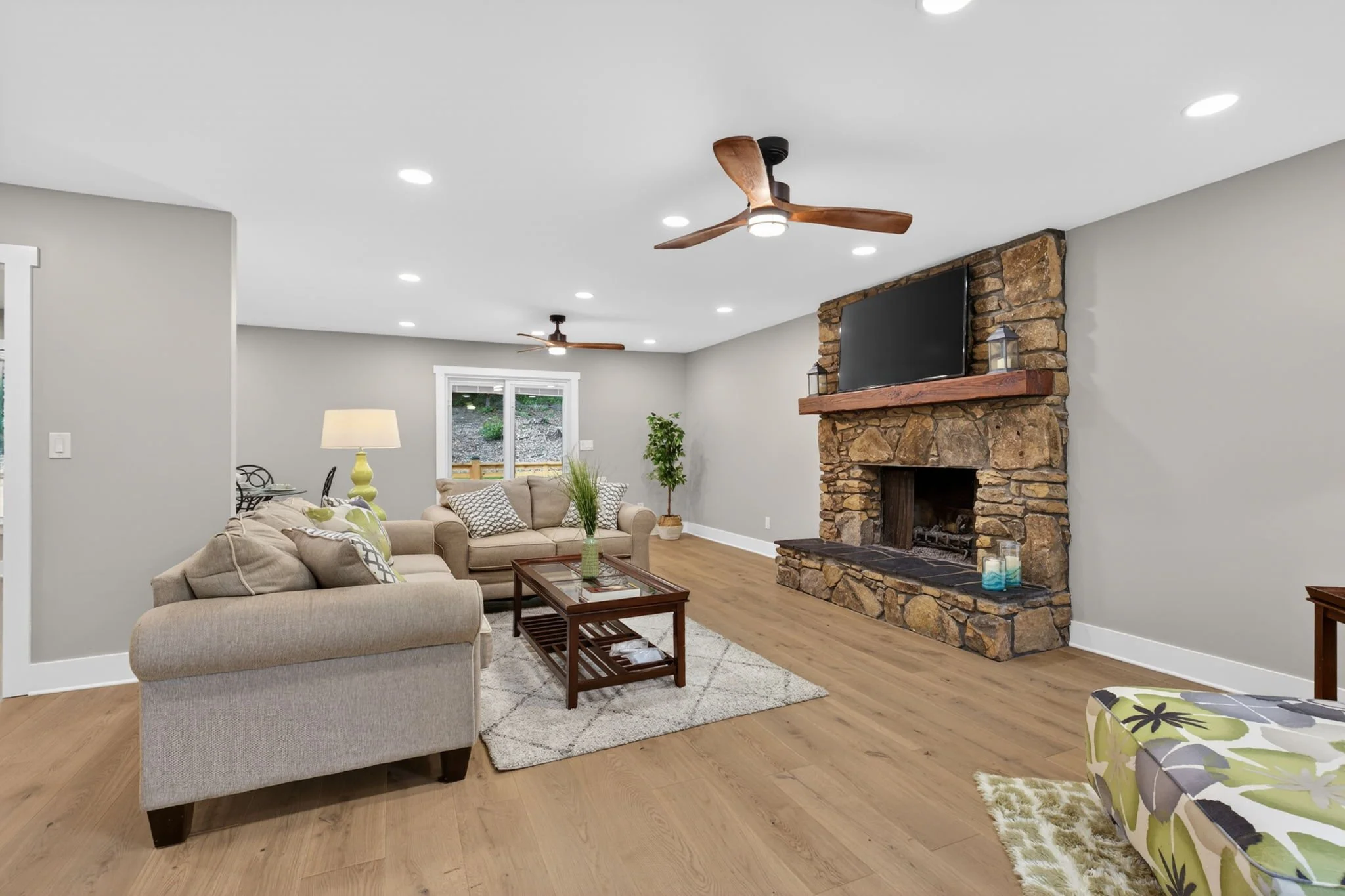 Living room with beige sofas, a stone fireplace with a mounted flat-screen TV, wooden floor, ceiling fans, a window showing a backyard, and a side table with a yellow lamp.