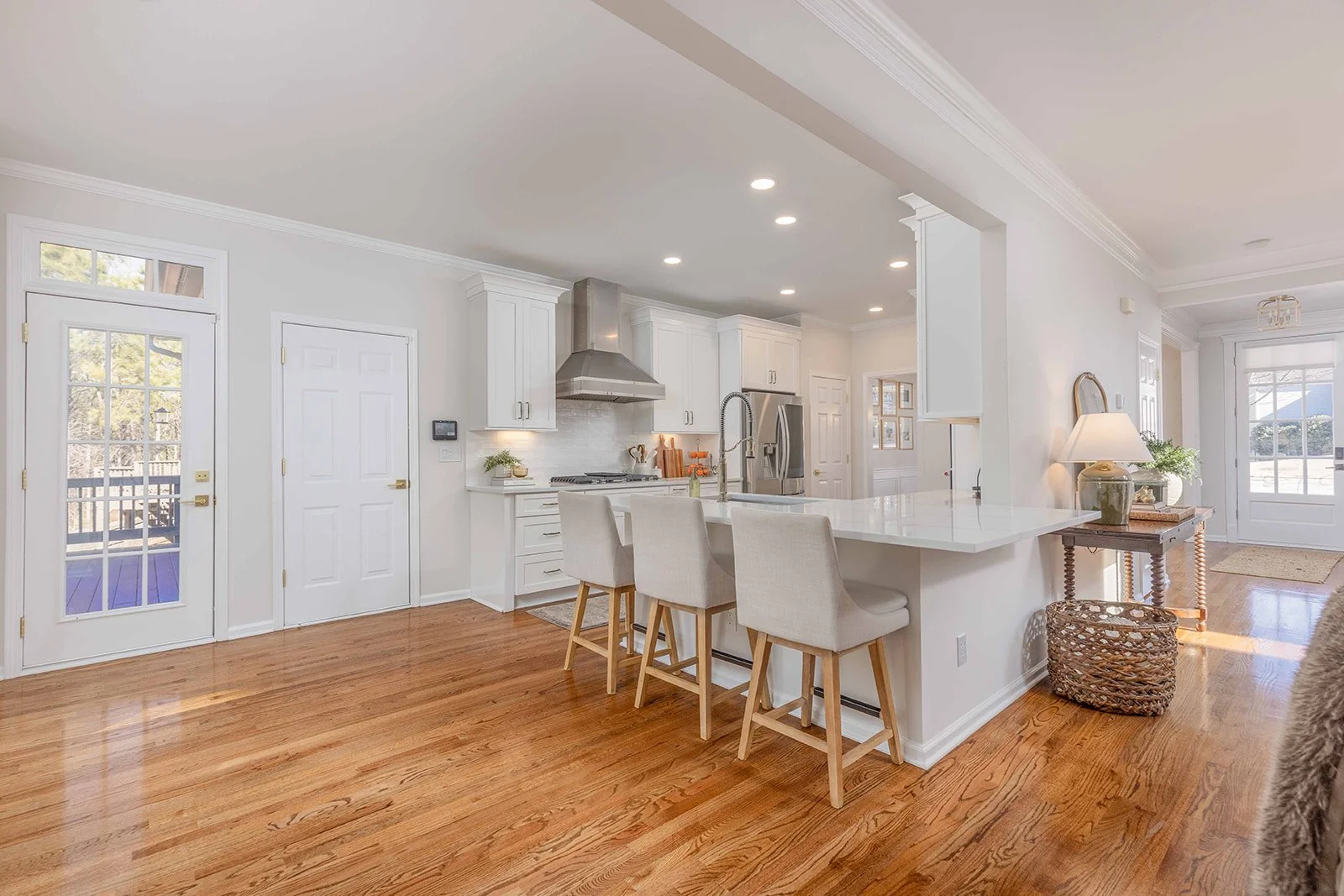 Bright, modern kitchen with white cabinets, stainless steel appliances, and a central island with four chairs. Hardwood floors, a door leading outside, and a small table with a lamp and plant near a window.