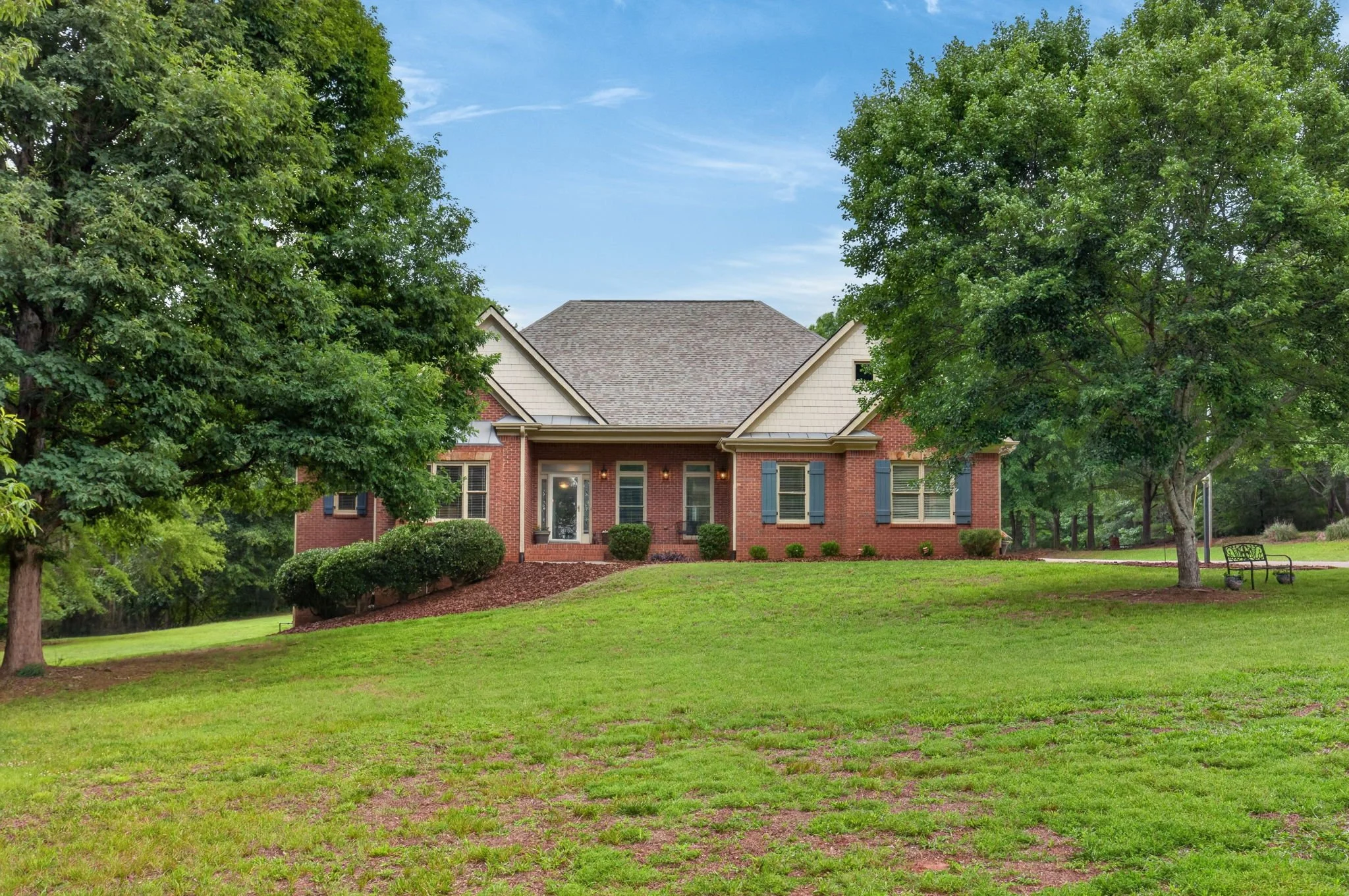 A brick house with a gray shingled roof surrounded by green trees and a well-maintained lawn.