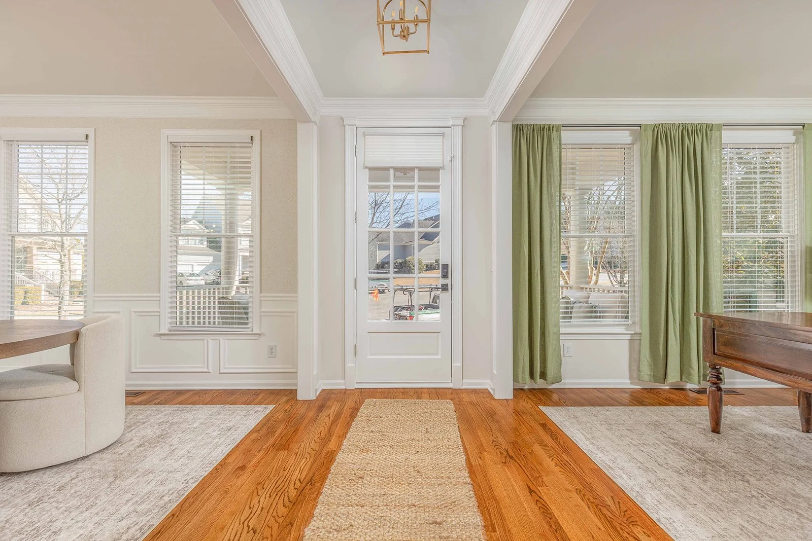 Bright entryway with white walls, hardwood floors, and multiple windows with blinds. Green curtains frame some windows, and a small rug runs from the door inward. Part of a round upholstered chair and a wooden table are visible.