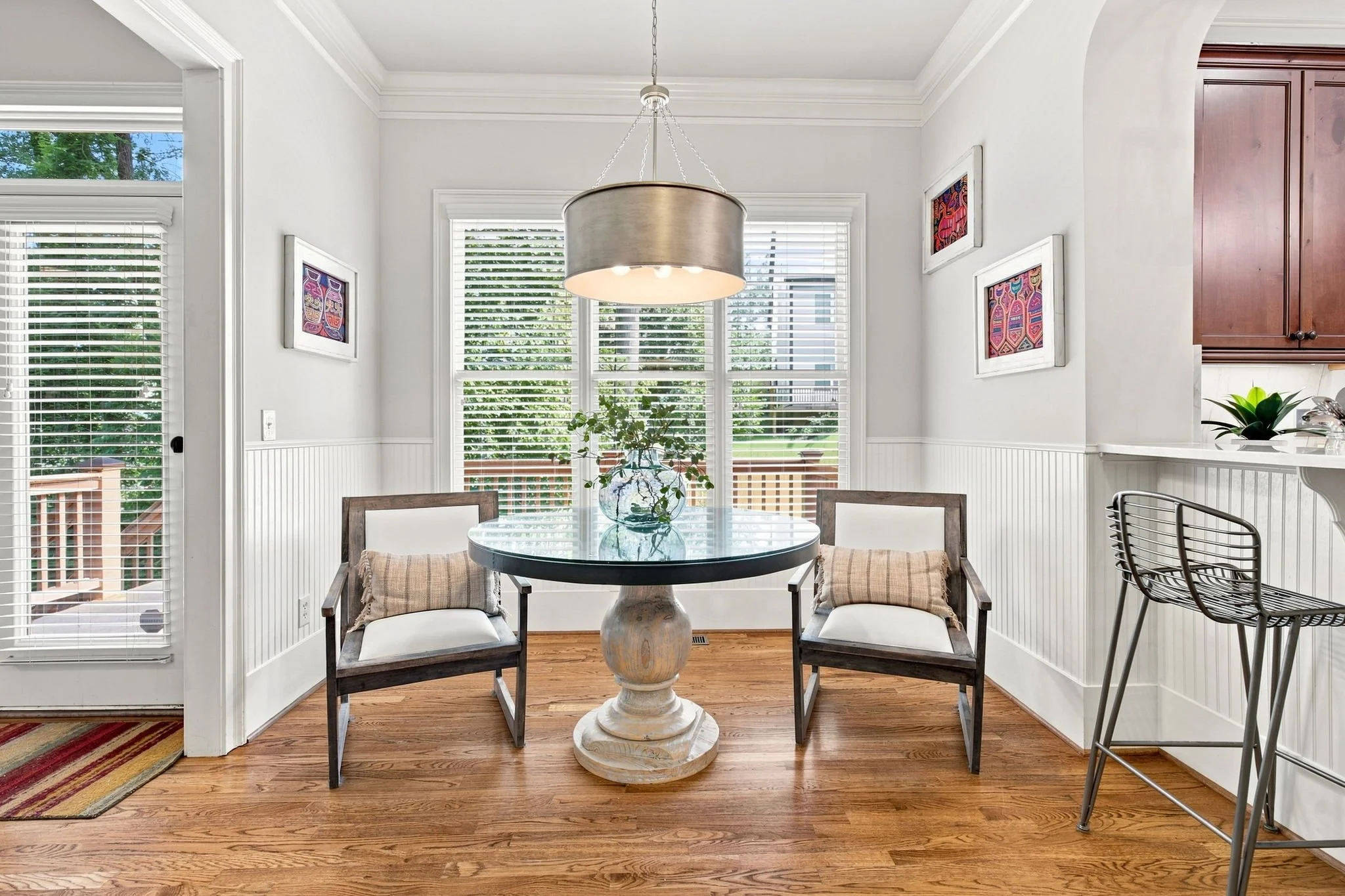 A dining area with a round glass-top table, surrounded by four chairs with cushioned seats, inside a room with white walls, large windows with white blinds, and hardwood floors. A pendant light hangs over the table, and a potted plant is on the table