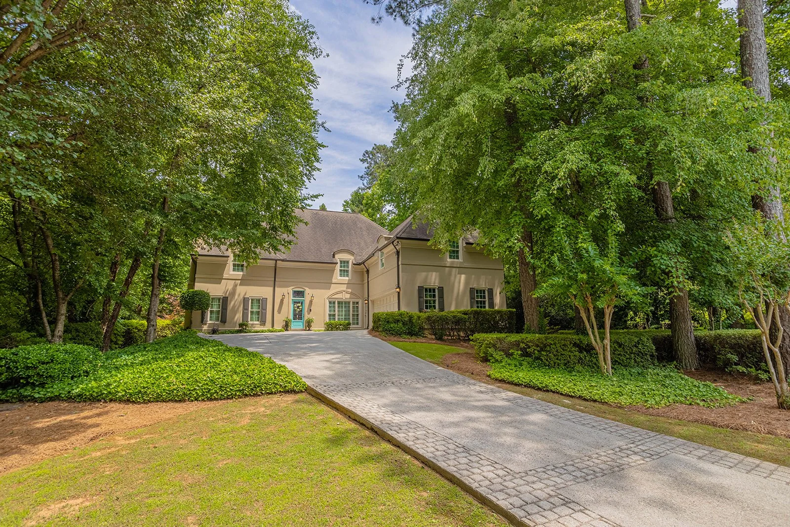Front view of a beige two-story house with a driveway surrounded by lush green trees and shrubs.