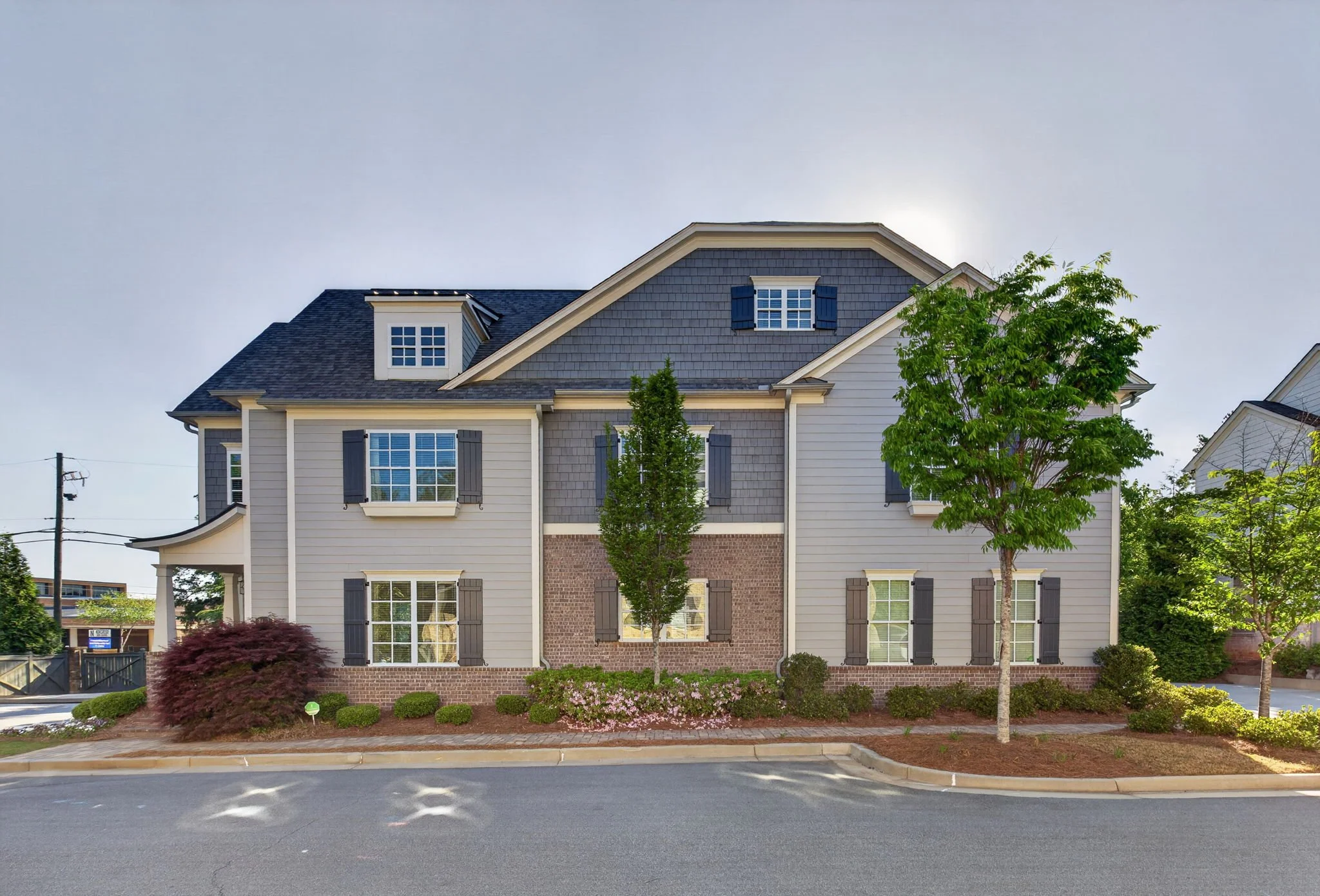 A multi-story house with gray siding, brick accents, and white trim, surrounded by neatly maintained landscaping including shrubs and trees, with a clear sky in the background.