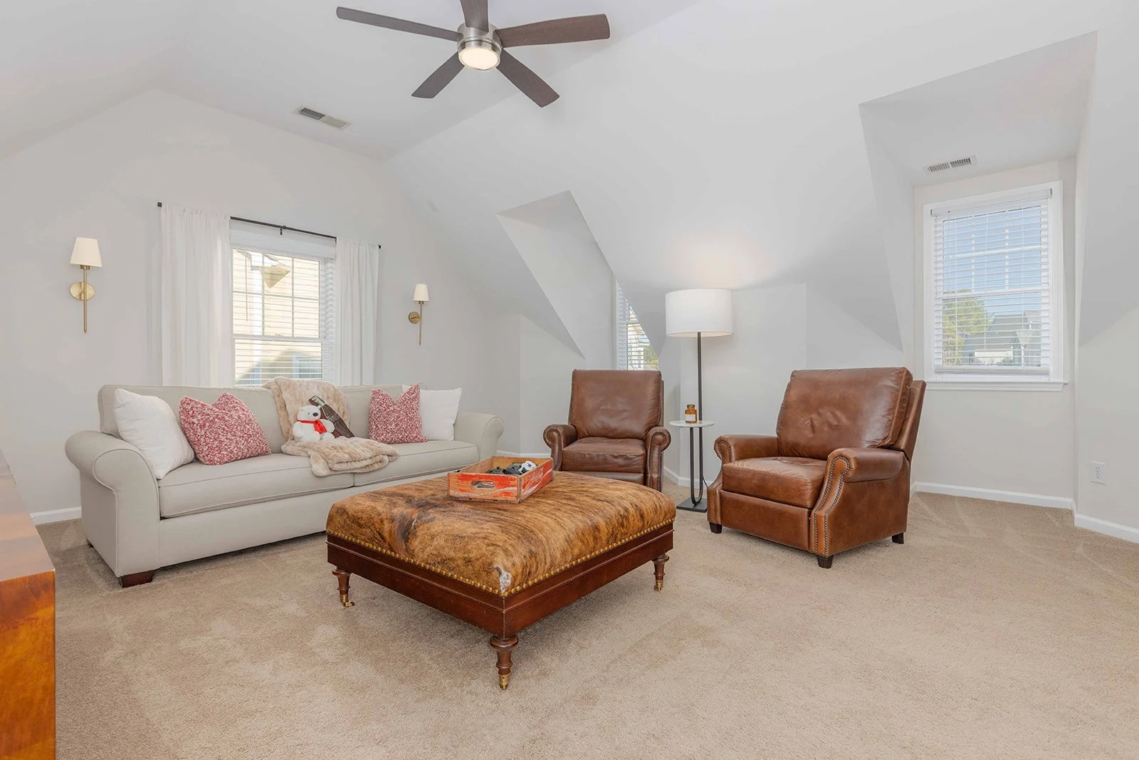 Living room with white walls, two brown leather armchairs, a white sofa with pink and plush pillows, a large brown ottoman, a ceiling fan, and windows with white blinds and curtains.