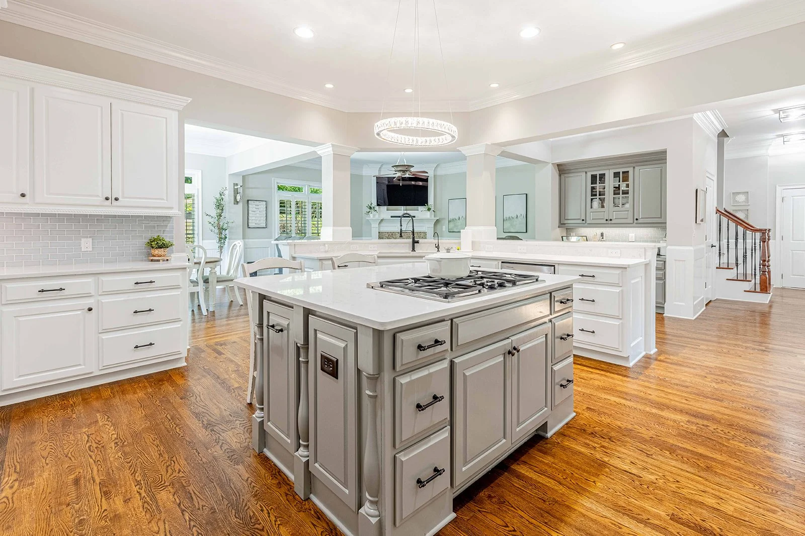 Open kitchen with white cabinets, a kitchen island with a grey base, hardwood floors, and a chandelier overhead. Brightly lit by recessed lighting and windows, with a dining area visible in the background.
