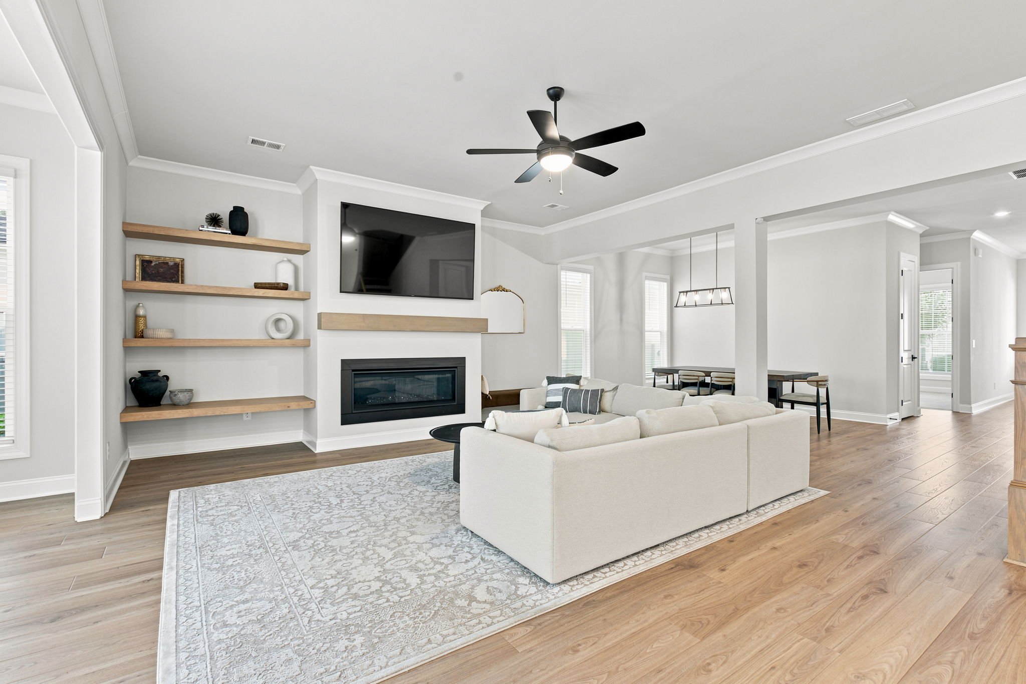 Living room with white walls, wooden floors, a large white sofa, a fireplace, a mounted flat-screen TV, wooden floating shelves with decor, and windows with blinds.