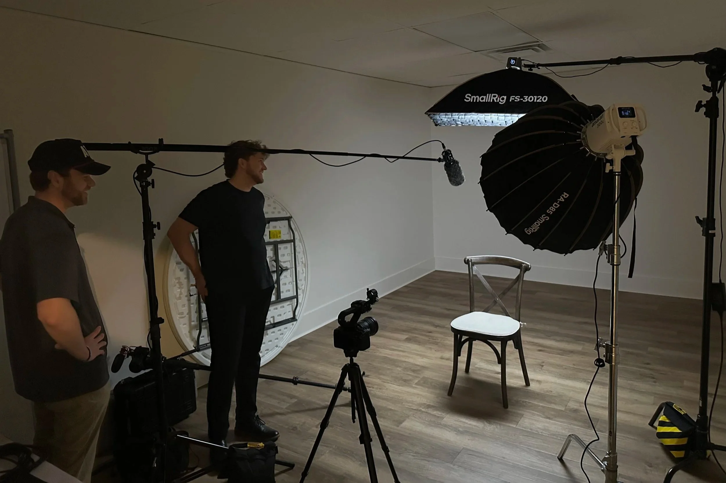 Two men in a photography studio with professional lighting and equipment, including a softbox and a reflector, around a simple chair.