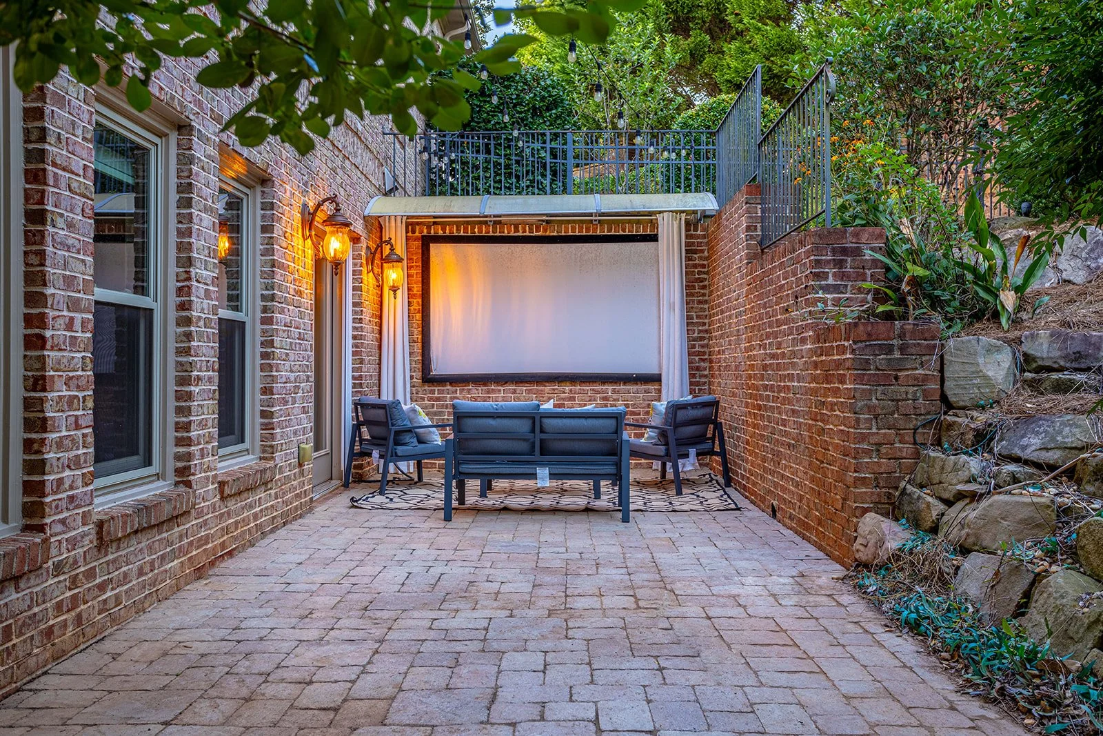 Outdoor patio area with brick walls, brick paving, a large screen for outdoor movies, and seating including chairs and a small table. There are string lights overhead and garden plants on the right side.