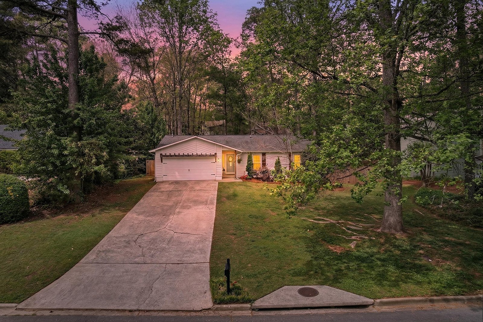 A single-story white house with a gray roof and attached garage, situated on a driveway. The front yard has grass and a large tree, with suburban neighborhood surroundings and a colorful sunset sky in the background.