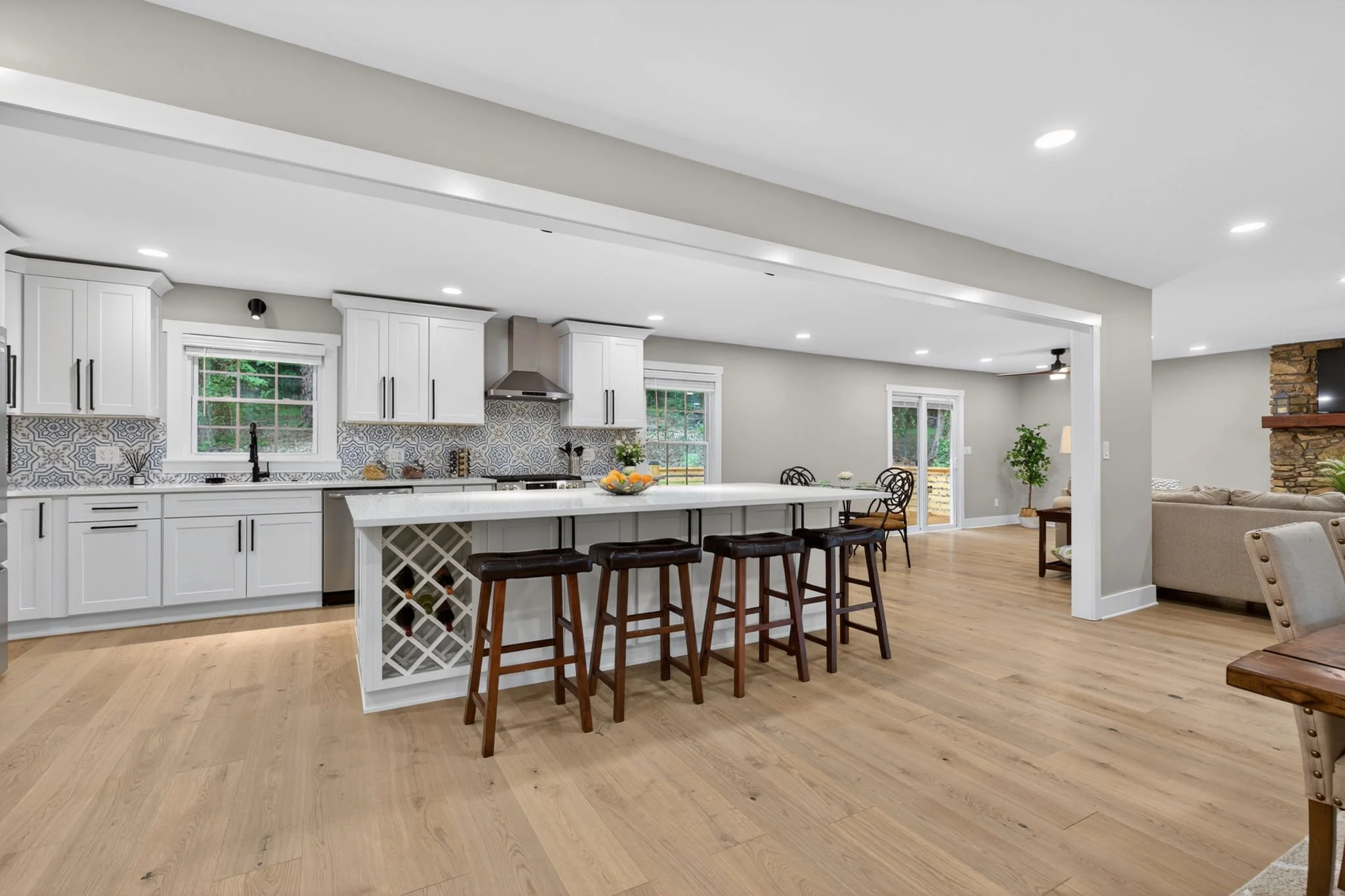 A modern, open-concept kitchen and living space with white cabinets, a patterned backsplash, a large island with bar stools, and wooden flooring.