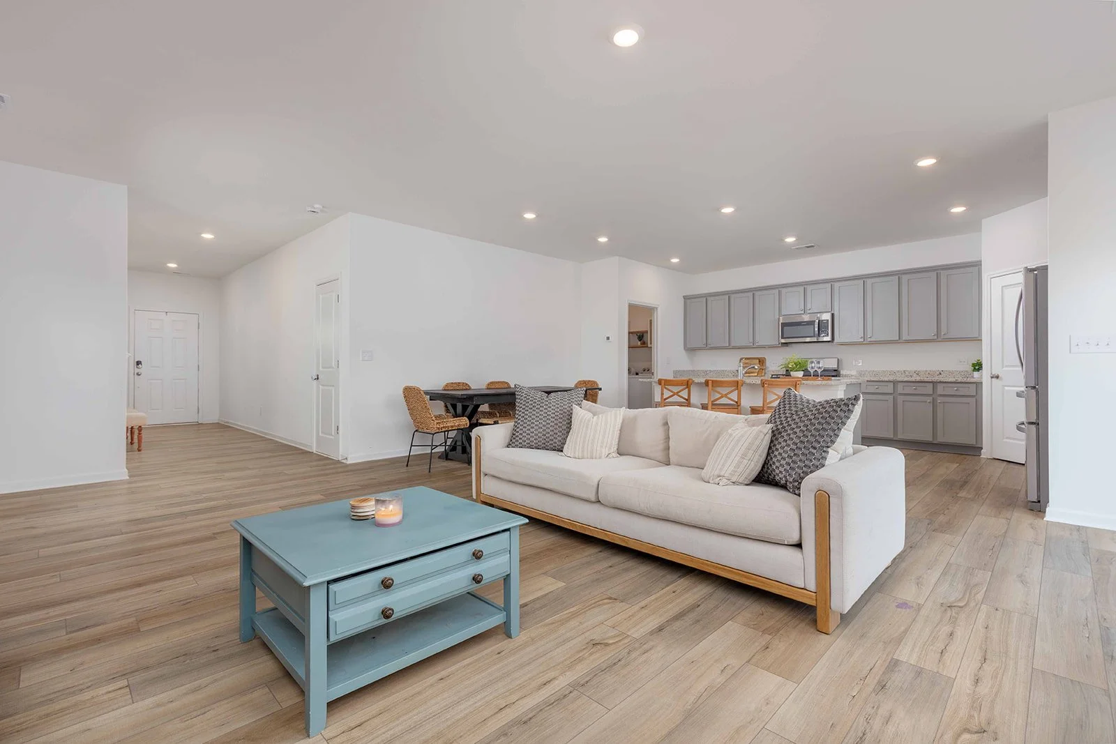 Open-concept living room and kitchen with white walls, light wood flooring, a cream sofa with pillows, a blue coffee table with candles, a dining table with wicker chairs, gray kitchen cabinets, and stainless steel appliances.