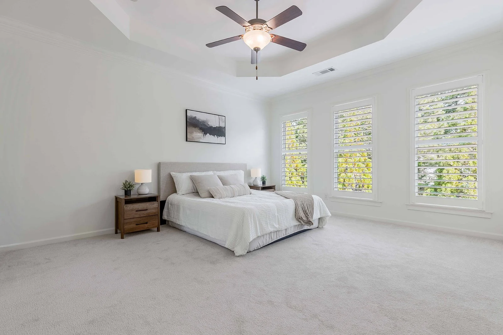A bright, minimalistic bedroom with white walls, a ceiling fan, a bed with white bedding and decorative pillows, side tables with lamps, and three large windows with shutters showing a view of trees outside.