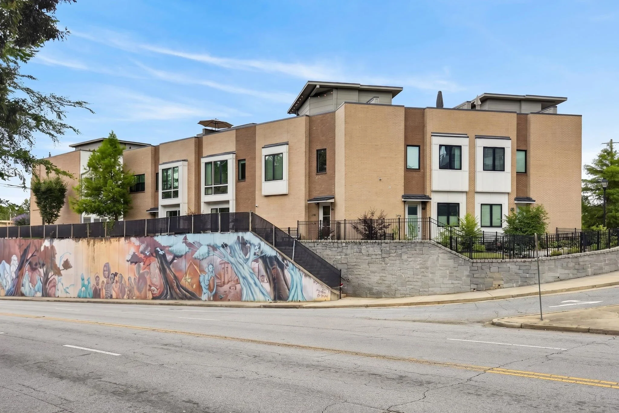 Modern multi-story residential building with beige and white exterior, large windows, and small rooftop balconies. There is a mural on a retaining wall in the foreground depicting a tree and people, with a street and sidewalk along the front of the b