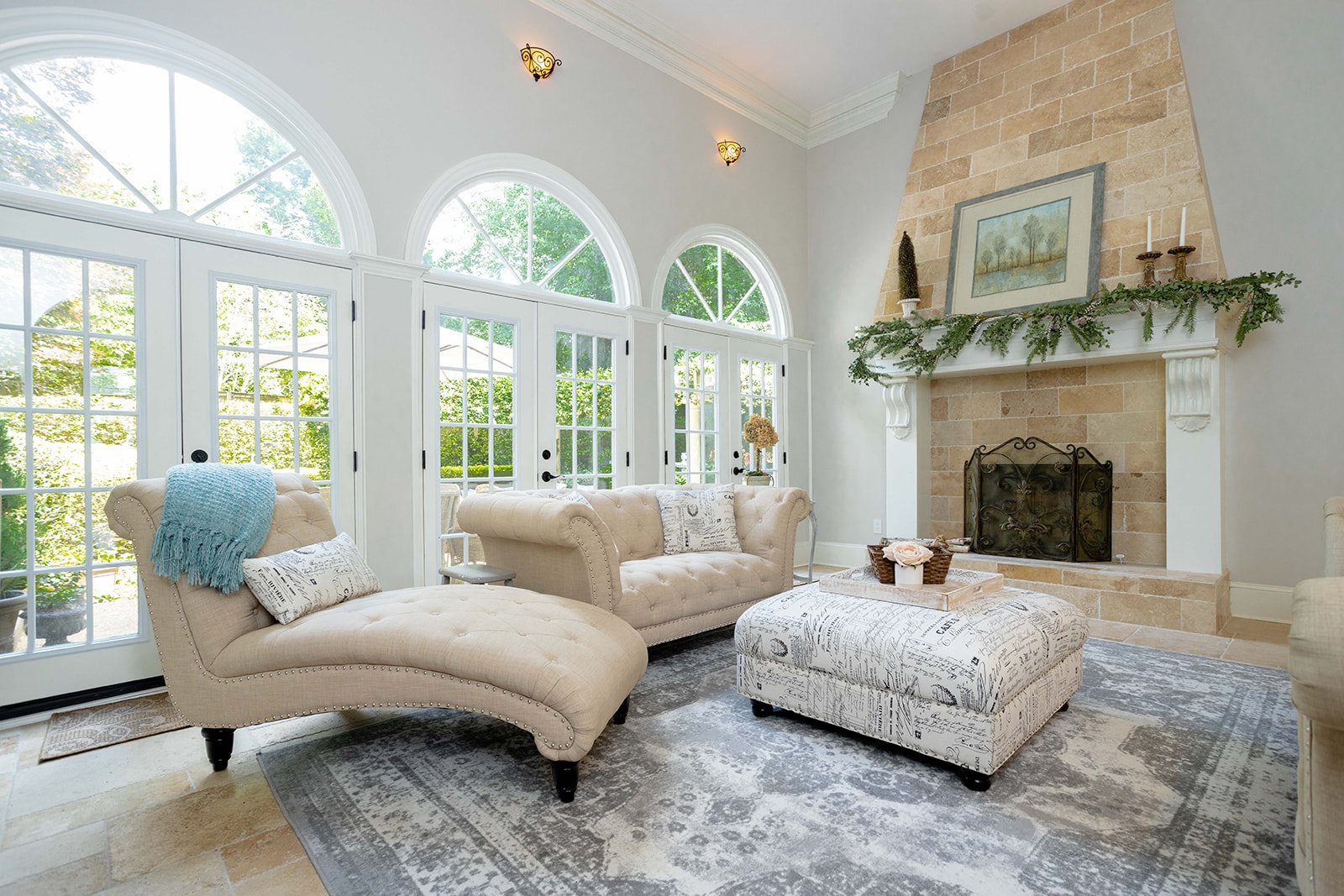 Bright living room with large arched windows, beige tufted sofas, a patterned ottoman, a stone fireplace with greenery and candles on mantel, and a decorative rug.
