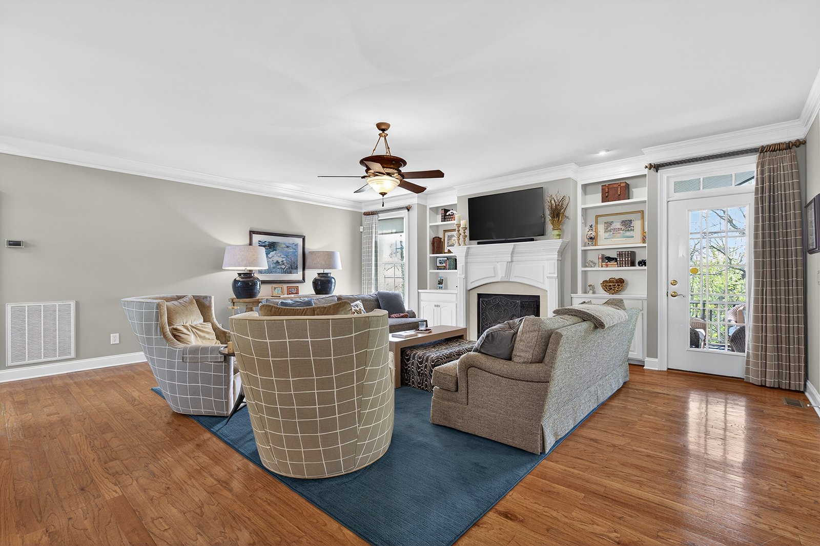 Living room with beige and gray furniture, hardwood floors, fireplace, built-in shelves, and a door leading outside. There are two lamps on side tables, a ceiling fan, and a wall-mounted TV above the fireplace.