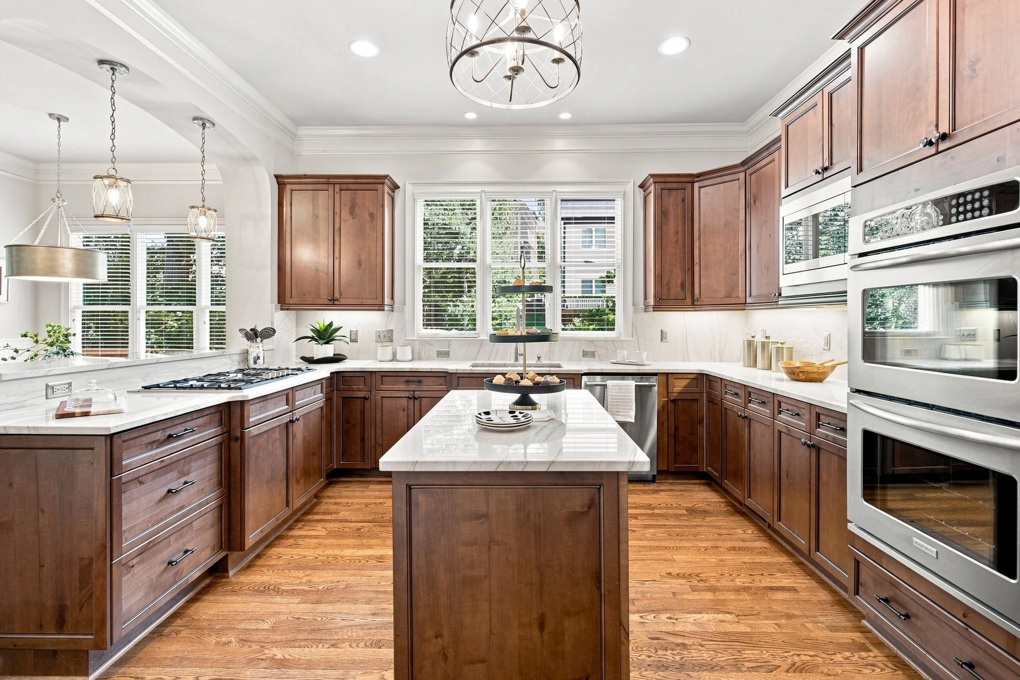 Bright kitchen with white marble countertops, wooden cabinets, multiple windows with white blinds, and stainless steel appliances. There are pendant lights hanging over the counter and a central island with a tiered serving stand with pastries.