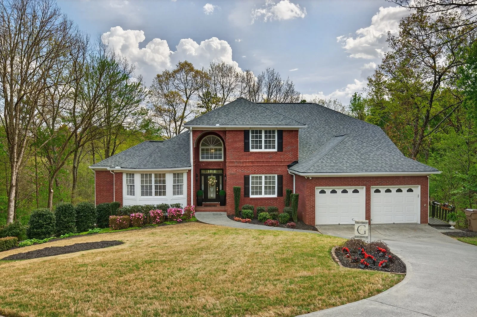 A two-story brick house with a gray shingled roof, white window trims, and black shutters. It has a landscaped front yard with pink flowers, green shrubs, and a curved concrete driveway leading to a two-car garage. There are trees behind the house an