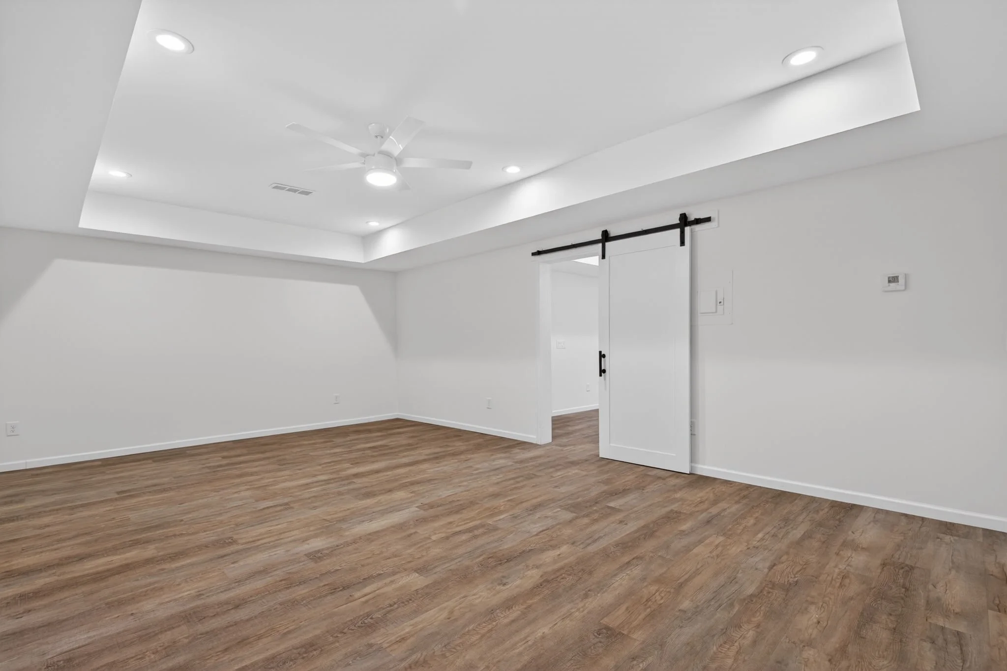 Empty room with white walls, a wooden floor, and a white ceiling with recessed lighting and a ceiling fan. There is a sliding barn door on a black track and a thermostat on the wall.