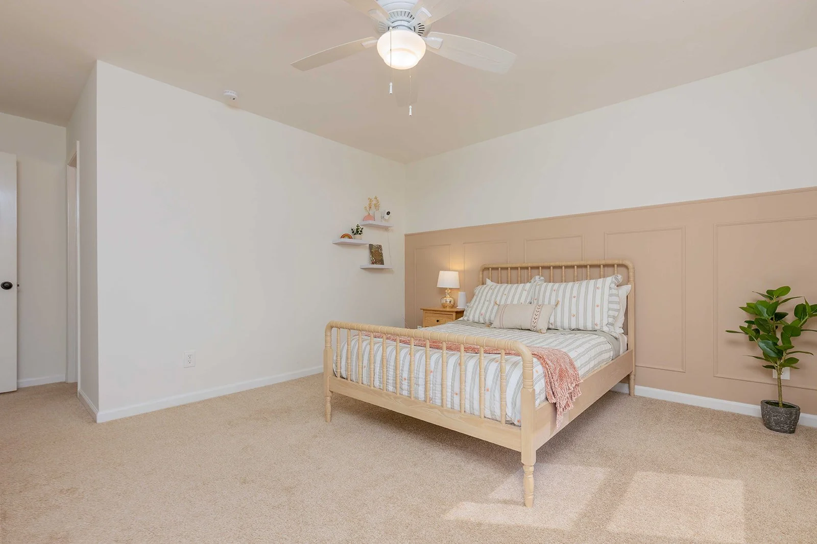 A bedroom with a bed, nightstand, lamp, and plant, featuring neutral tones and wall decor.