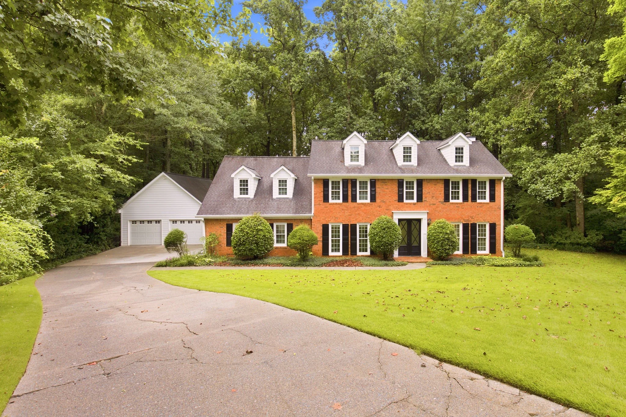 A brick two-story house with white-trimmed windows and black shutters, surrounded by green bushes and trees, with a curved driveway leading to a garage on the left, and a lush green lawn in the front.