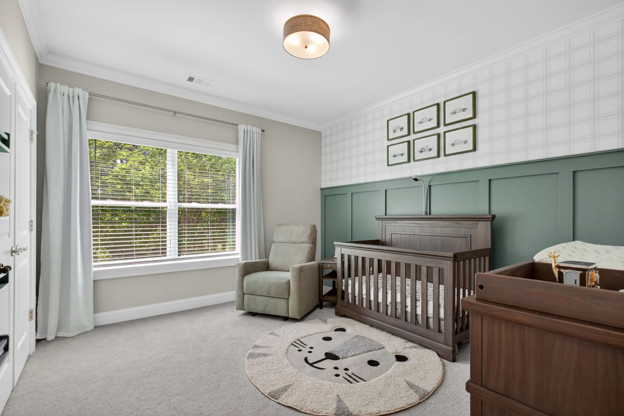 A cozy nursery with a large window featuring white blinds and curtains, a beige armchair, a wooden crib, a chest of drawers, and a round rug with a smiling cat face. Art prints of cars are hung on a green half-wall.