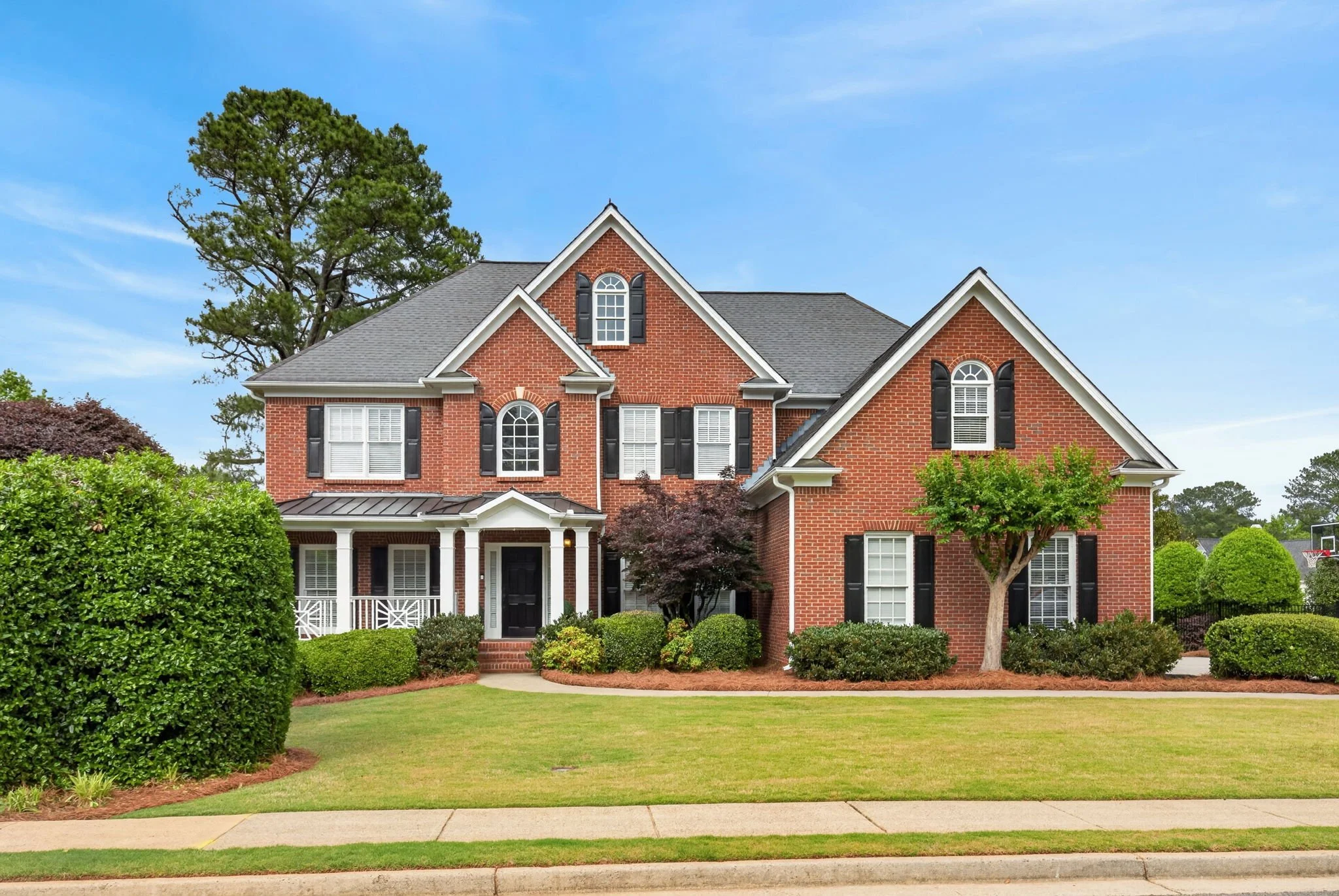 A two-story red brick house with white trim and black shutters, surrounded by green trees and shrubs, with a lawn and sidewalk in the foreground and a blue sky overhead.