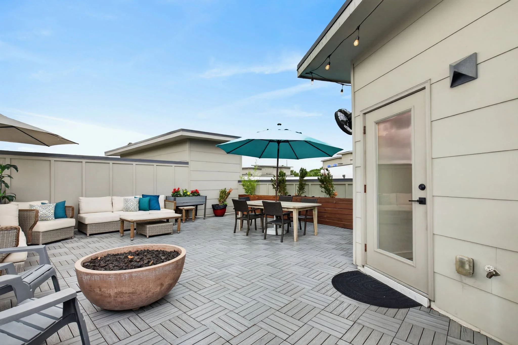 An outdoor patio area with a white sectional sofa, a round flower pot, several tables with chairs, umbrellas, and a beige fence under a blue sky.
