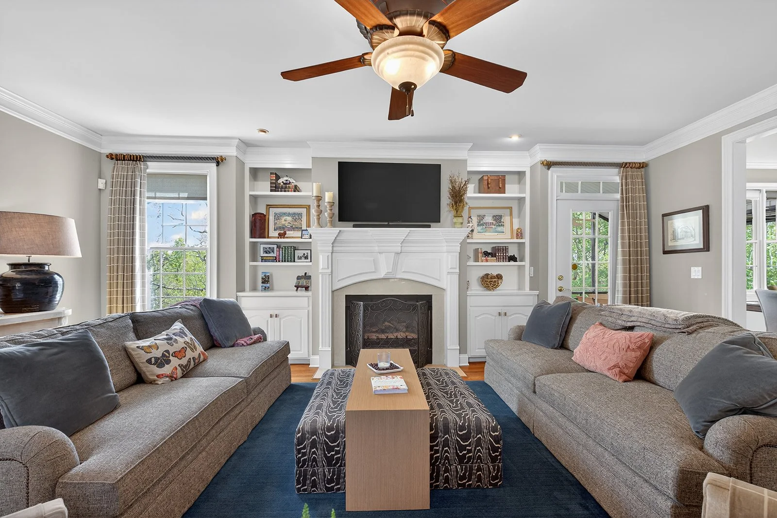 A cozy living room featuring two beige sofas with patterned and plain pillows, a wooden coffee table with magazines and candles, a white fireplace with built-in shelves, a wall-mounted TV above the fireplace, and windows with plaid curtains letting i