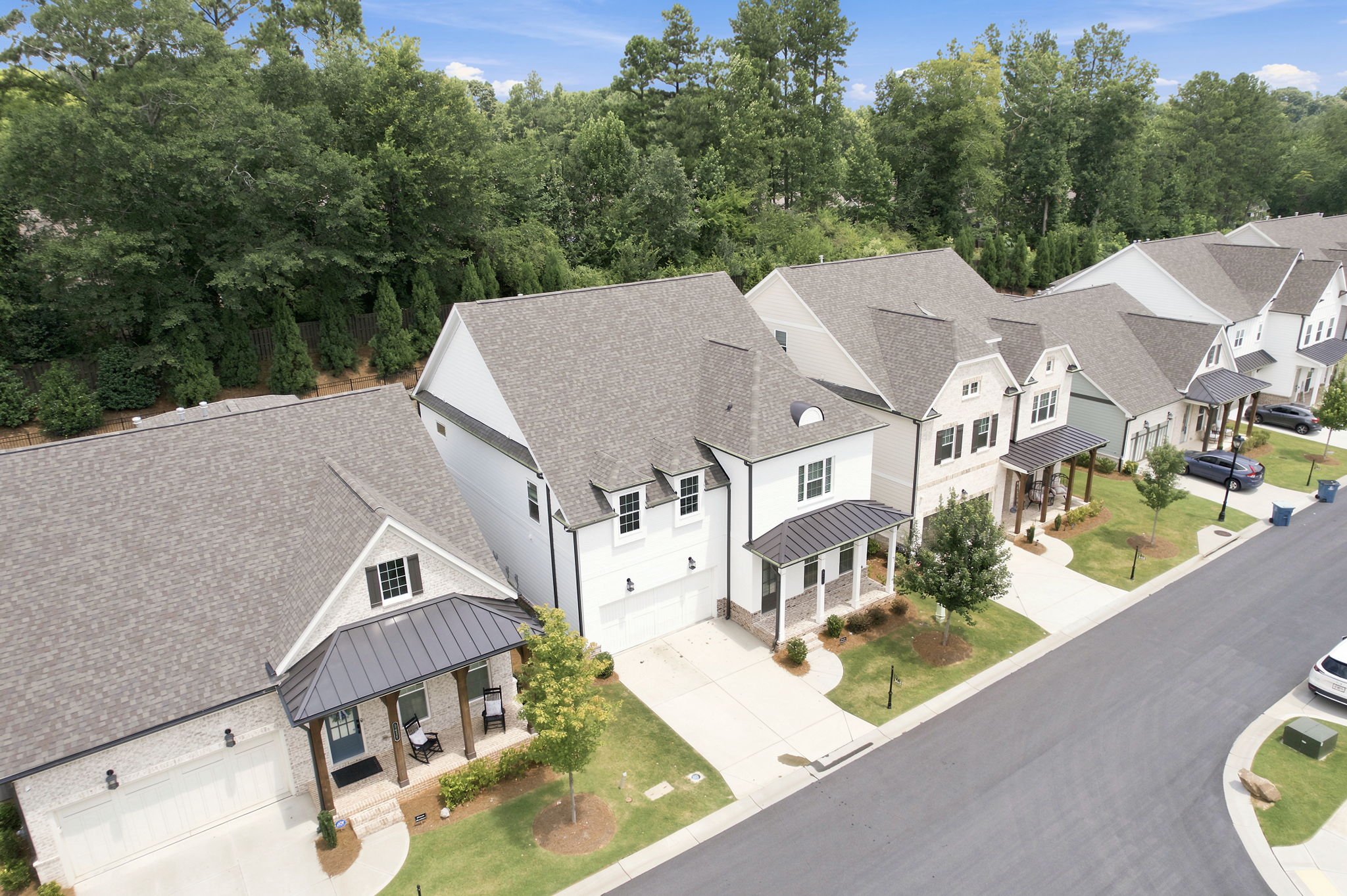 Aerial view of suburban neighborhood with white houses, gray roofs, driveways, parked cars, green lawns, and a wooded area in the background under a partly cloudy sky.