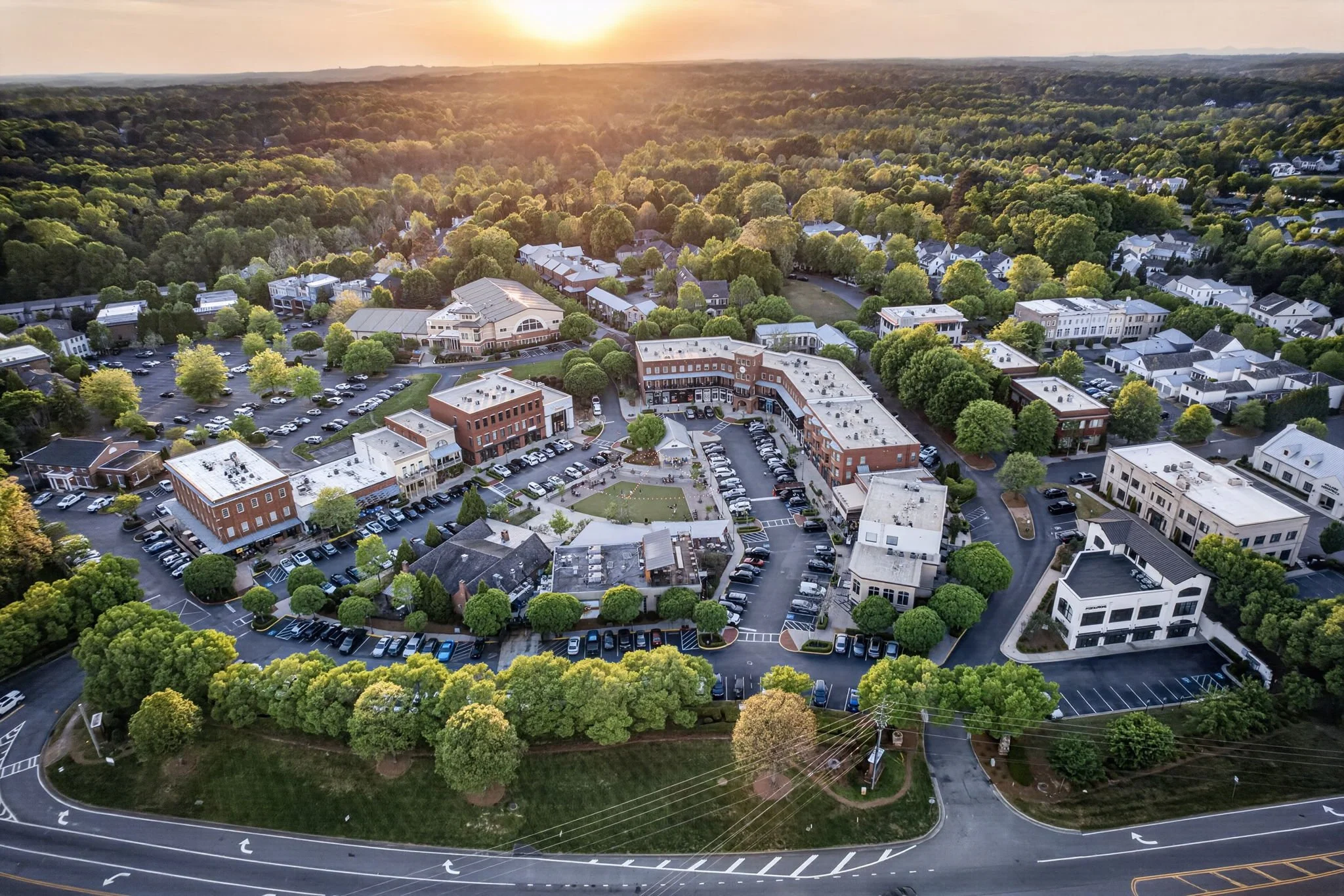An aerial view of a suburban neighborhood at sunset with a mix of single-family homes, commercial buildings, parking lots, and lush green trees. The sun is low on the horizon, casting a warm glow over the area.