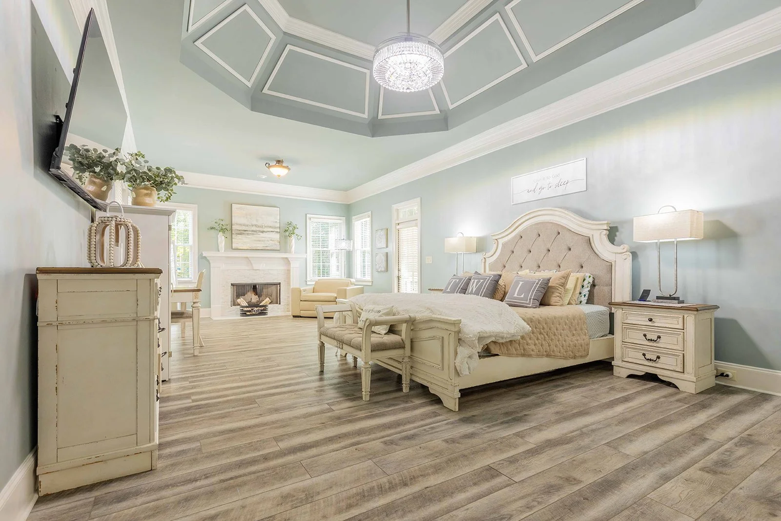Elegant bedroom with light blue walls, a large bed with a tufted beige headboard, matching nightstands with lamps, a fireplace, a beige armchair, and a ceiling with geometric molding and a chandelier.