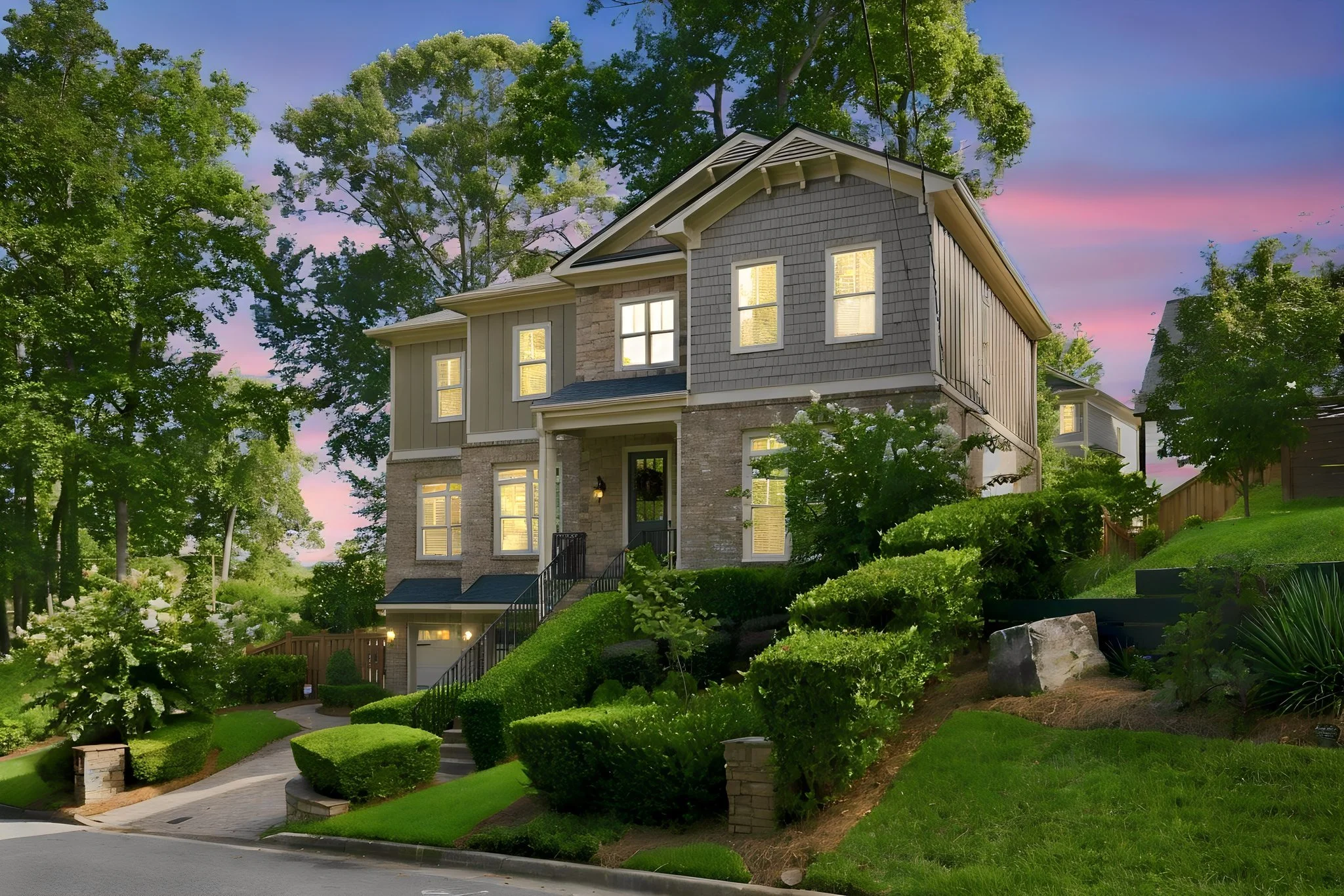 A multi-story house with a mix of brick and gray siding, lit from inside, surrounded by lush green trees and well-maintained landscaping, during the evening with a colorful sunset sky.