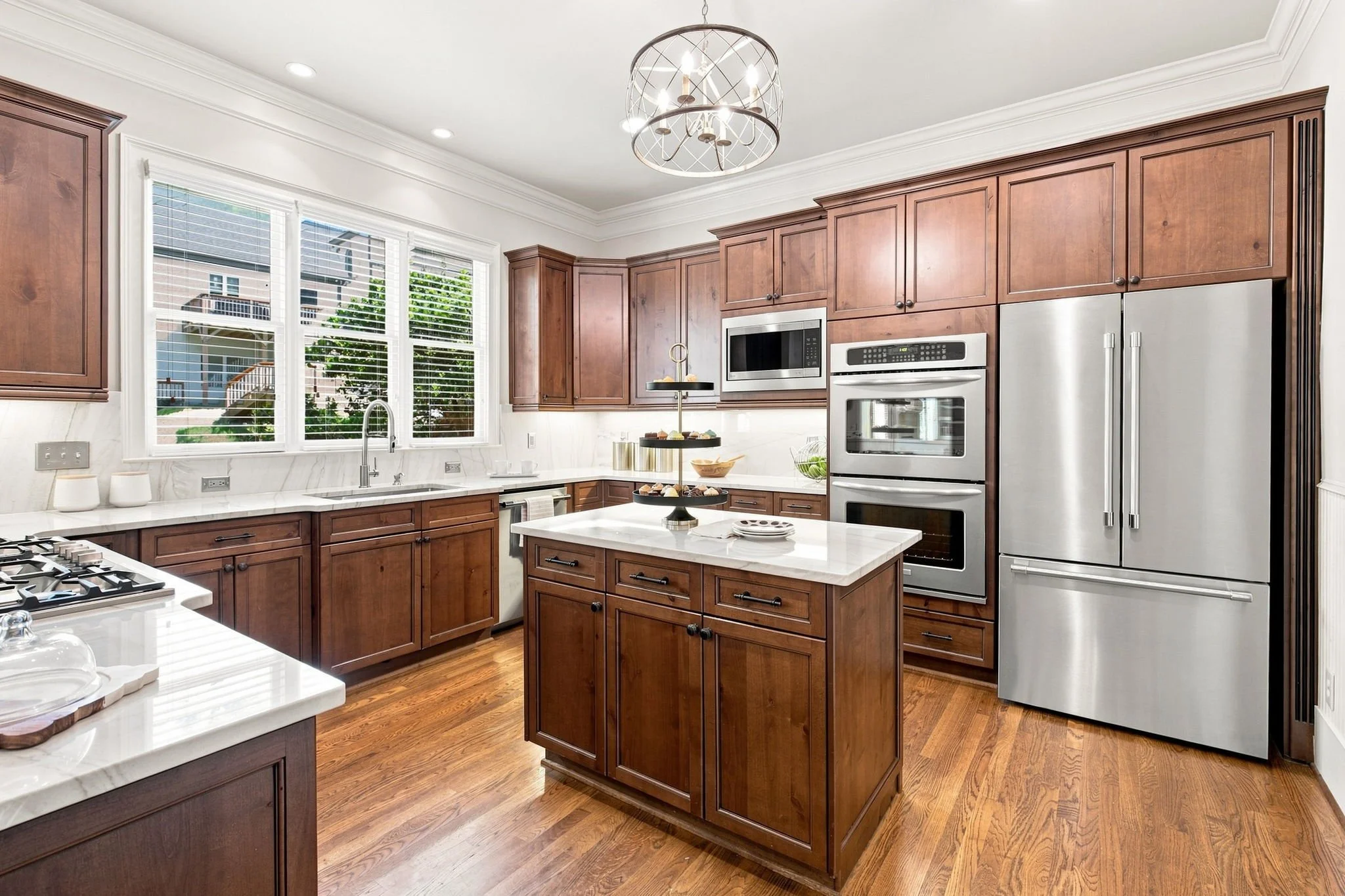 A modern kitchen with wooden cabinets, white marble countertops, large window with white blinds, stainless steel appliances including a double oven and microwave, and a white kitchen island with decorative cake stands and a bowl of fruit.