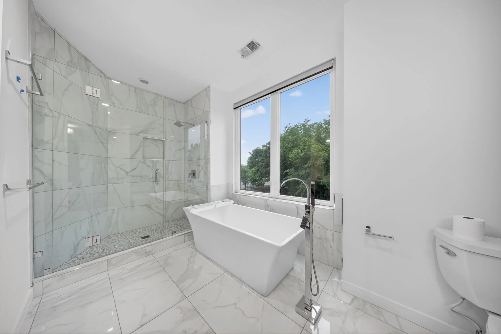 Modern bathroom with large window, marble shower area with glass door, freestanding bathtub, and white walls.
