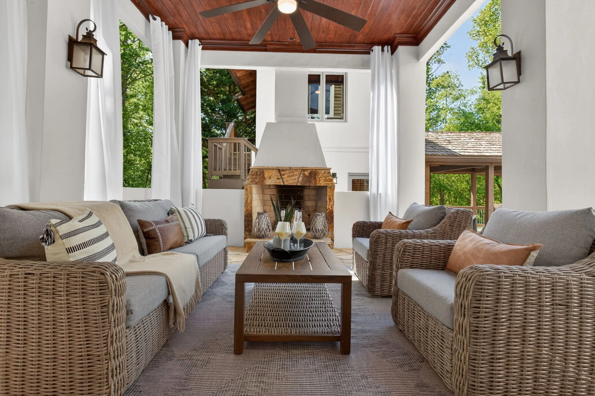A bright and airy outdoor living space with a wooden ceiling, ceiling fan, white curtains, and a stone fireplace. There are wicker sofas with cushions, a wooden coffee table, and trees visible outside the open areas.