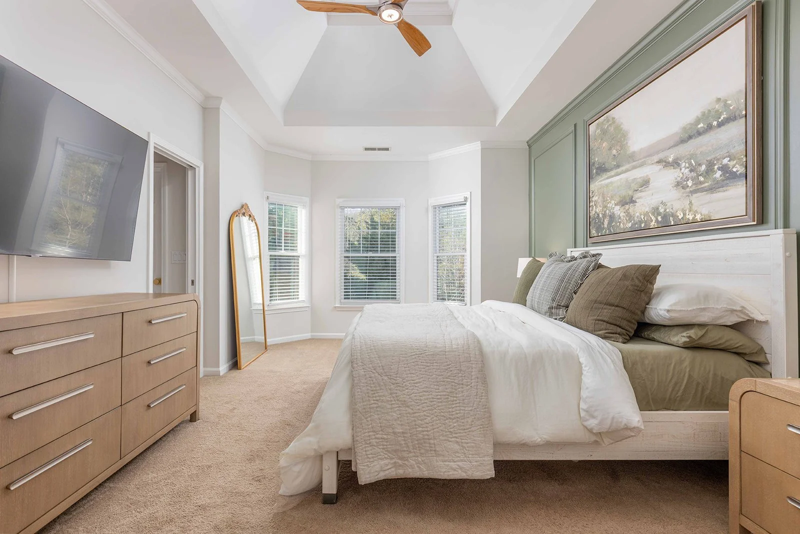 A bedroom with a large bed, seven pillows, white bedding, beige carpet, and a green accent wall with a landscape painting. There are three windows with white blinds, a wooden dresser with a TV on top, a tall mirror, a ceiling fan, and a door.