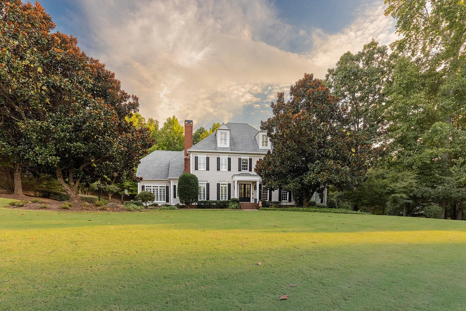 A large white house with black shutters, surrounded by trees and a well-maintained lawn, under a partly cloudy sky.