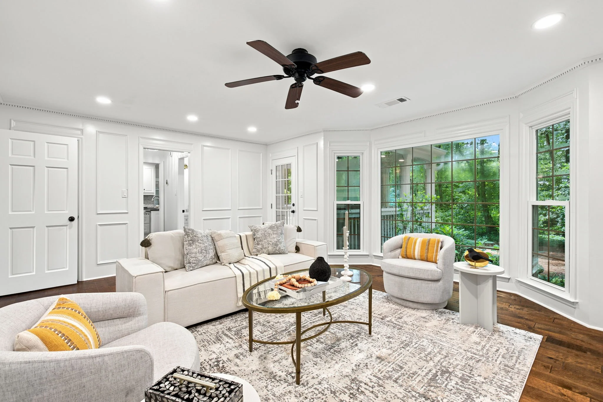 Living room with large windows showing trees outside, white walls with decorative molding, a ceiling fan, and white furniture including a sofa and armchair with yellow and gray pillows, a glass coffee table with decor items, and a patterned area rug.