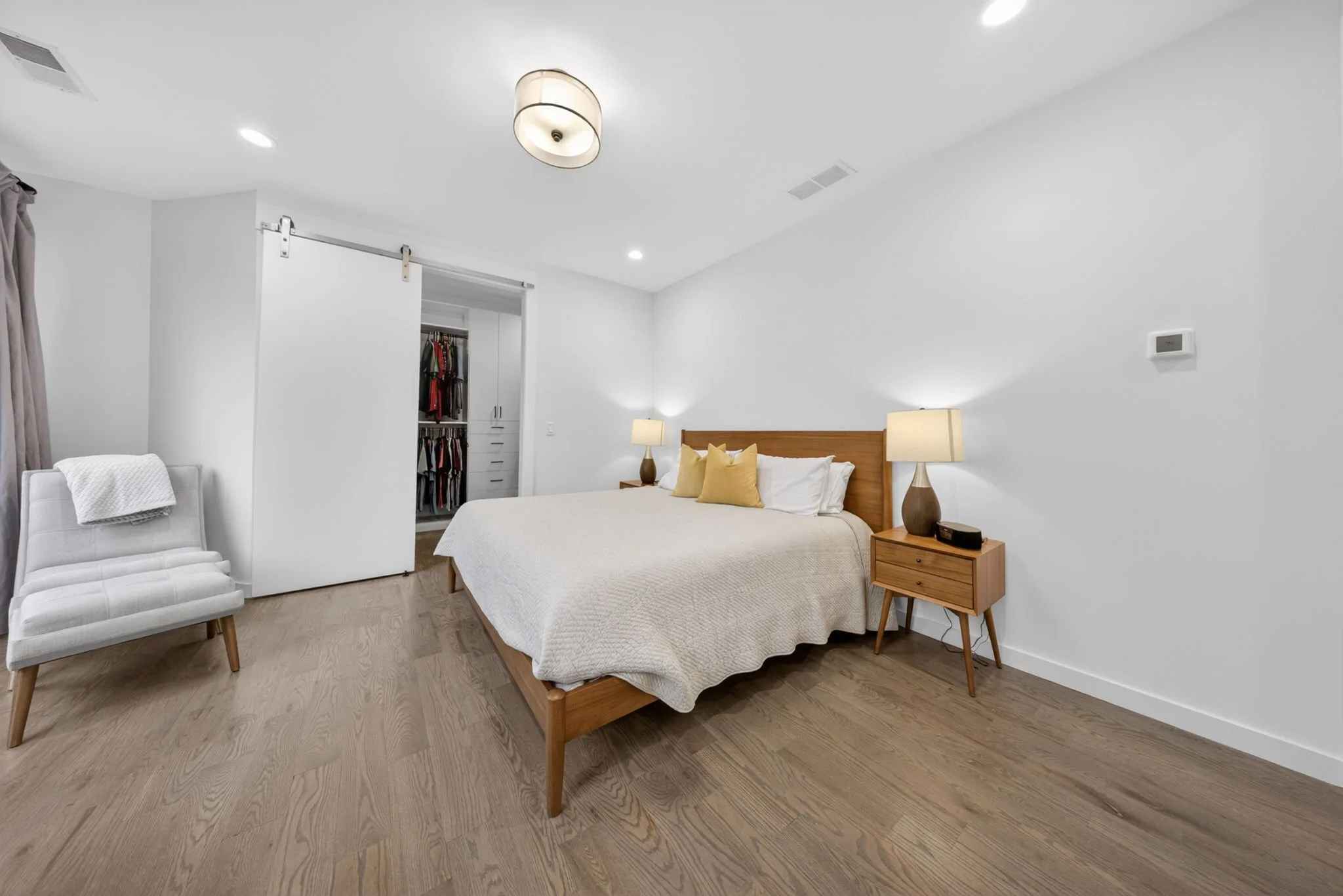 A bedroom with a wooden bed frame, beige bedding, and four pillows. Two bedside lamps on wooden nightstands. A white armchair with a throw blanket, an open closet with clothes, and a finished wooden floor.