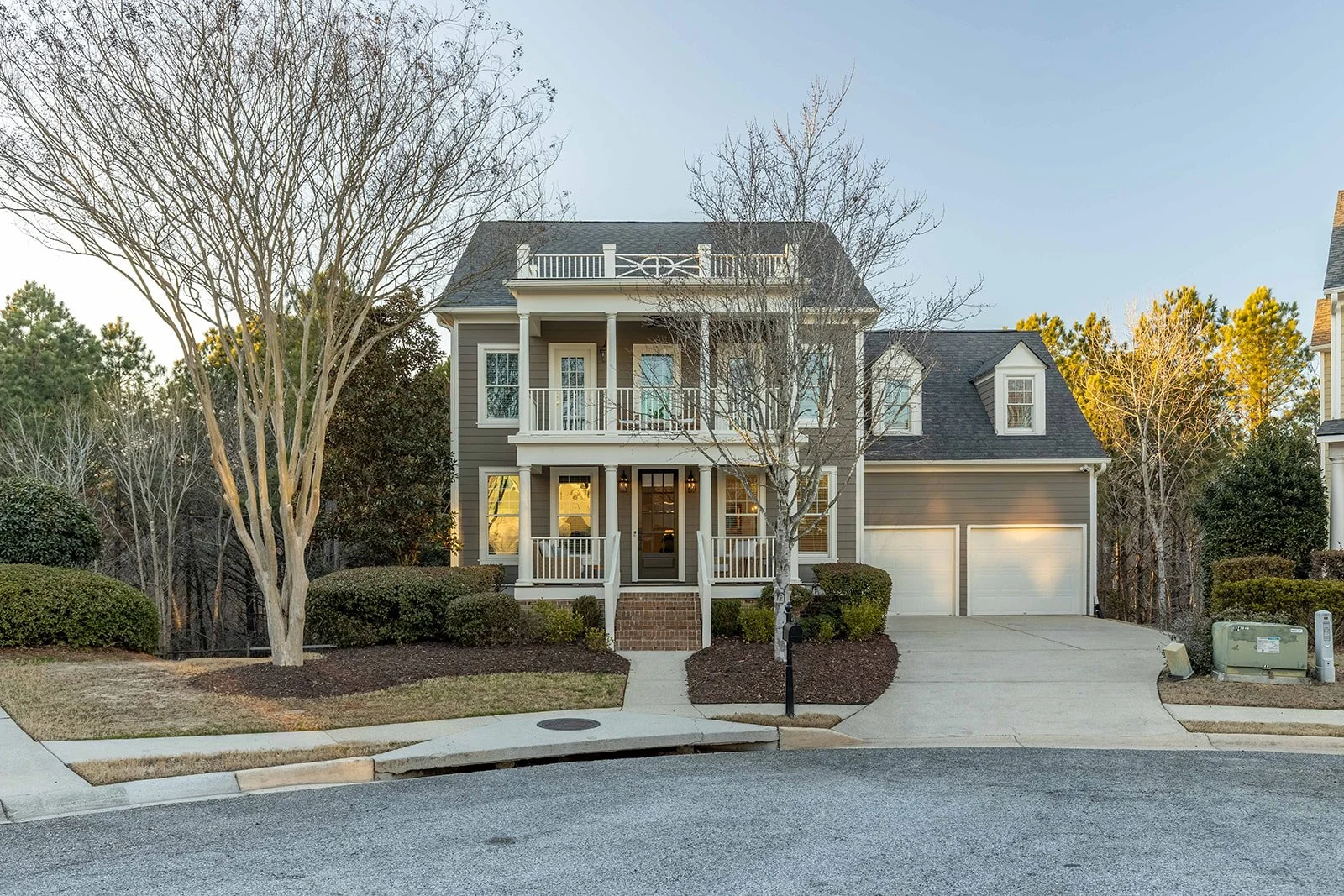A two-story house with a covered front porch, gray siding, white trim, and a two-car garage. There are leafless trees and bushes in the front yard, and the driveway and sidewalk lead to the front steps.