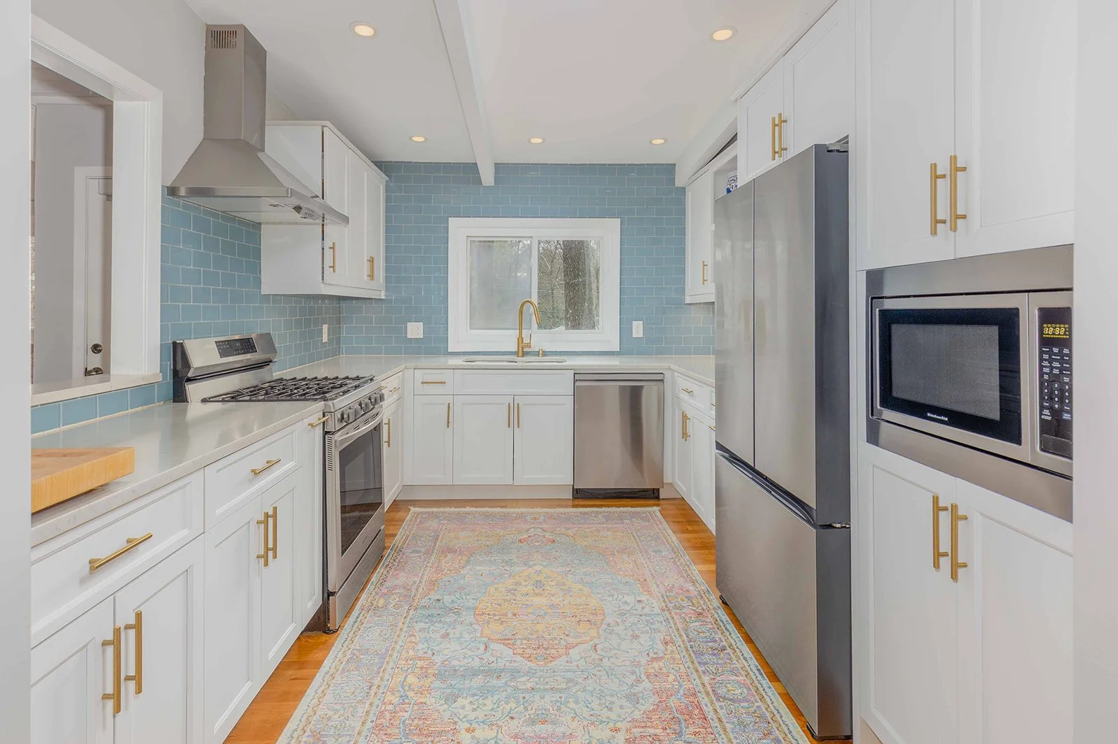 Modern kitchen with white cabinets, stainless steel appliances, light blue tile backsplash, a window, and a colorful rug on wooden floor.