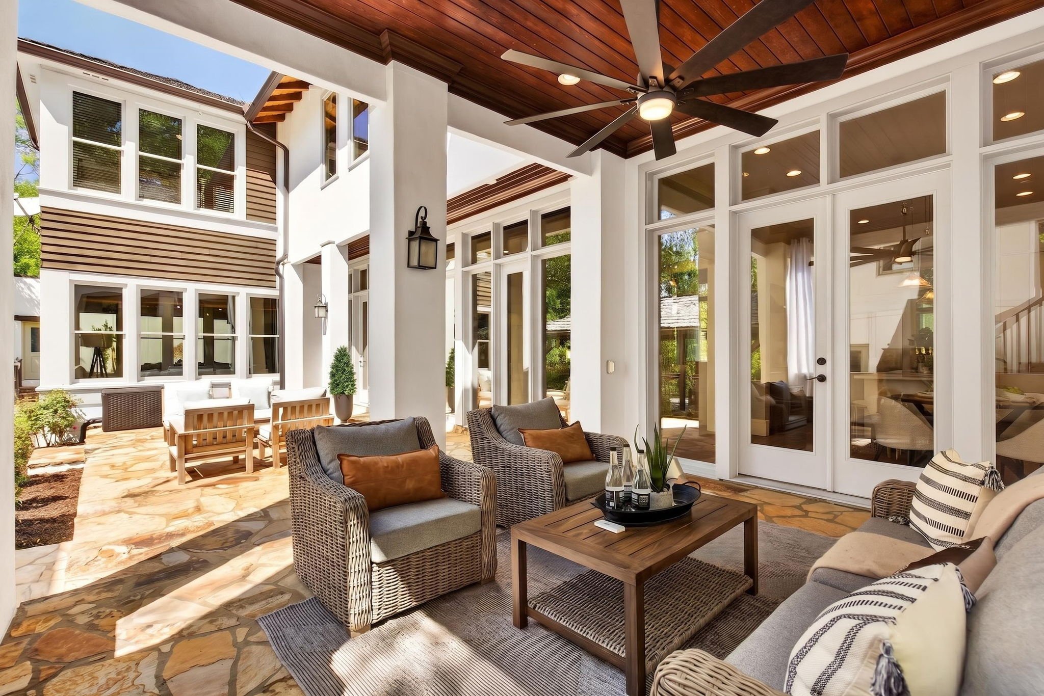 Covered patio area with wicker and cushioned seating, coffee table with bottles, ceiling fan, and large windows and doors to interior of house.