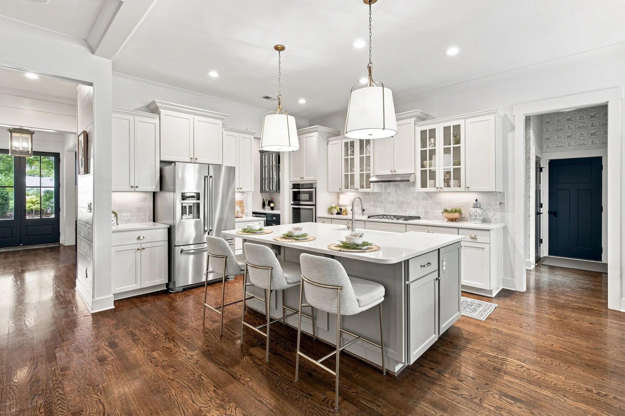 A modern kitchen with white cabinets, stainless steel appliances, a large white island with seating, dark hardwood flooring, and pendant lighting.