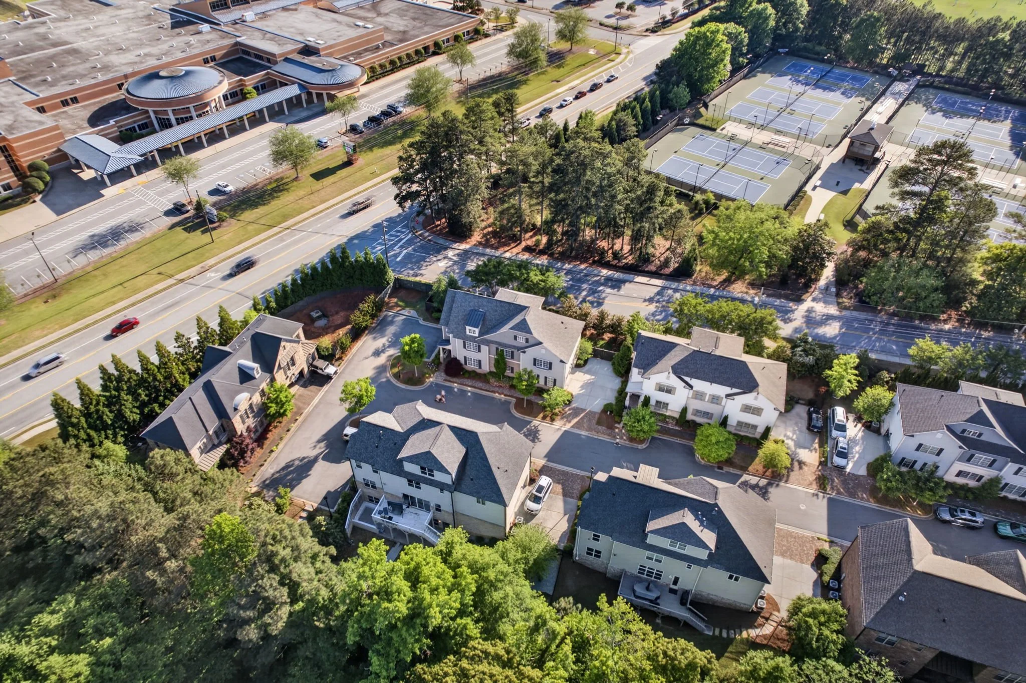Aerial view of a suburban area showing residential houses, a sports complex with tennis courts, a large building that appears to be a school or community center, and tree-lined streets with cars.