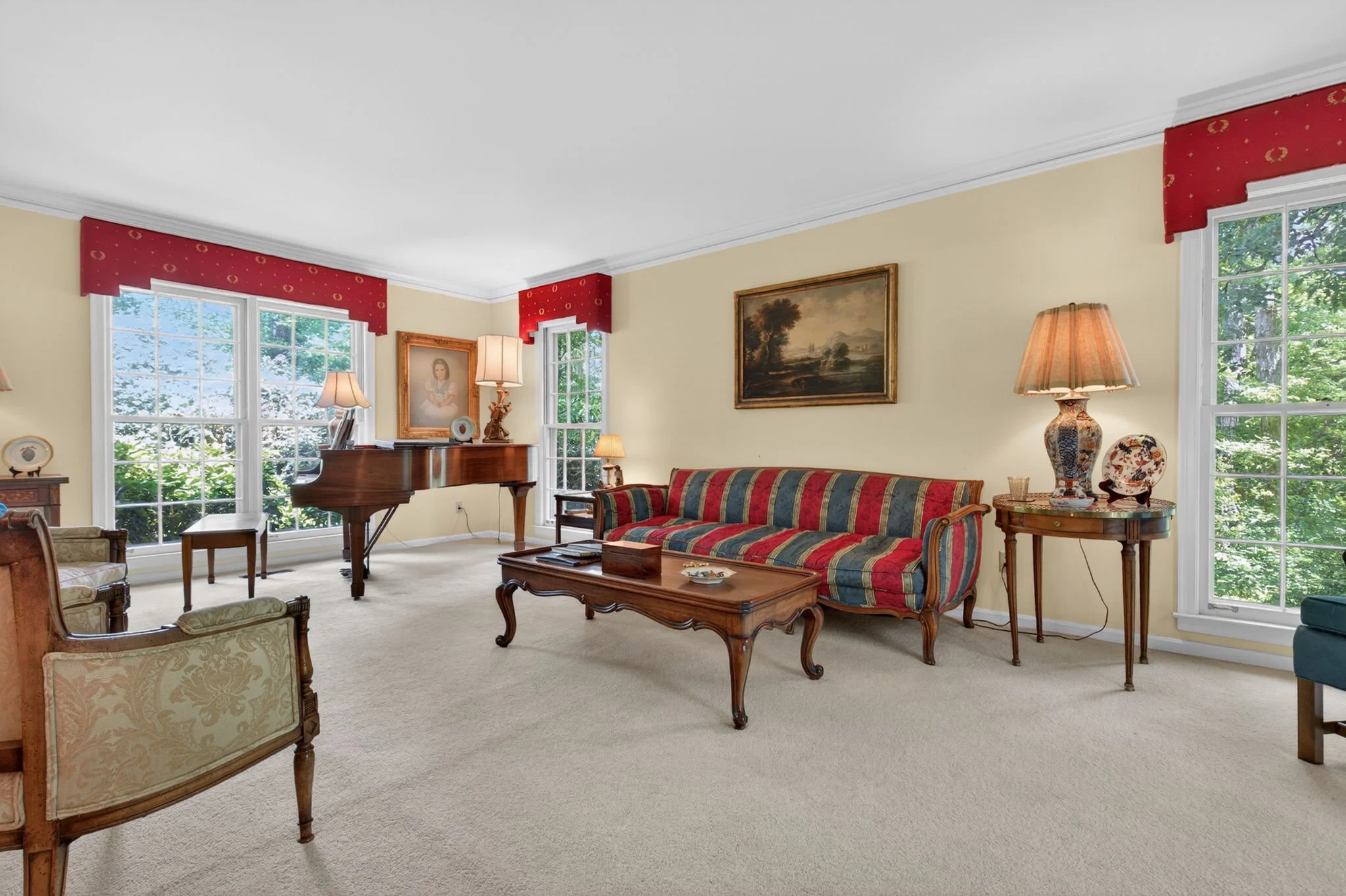 A living room with large windows, a grand piano, a striped sofa, framed paintings, lamps, and traditional furniture.