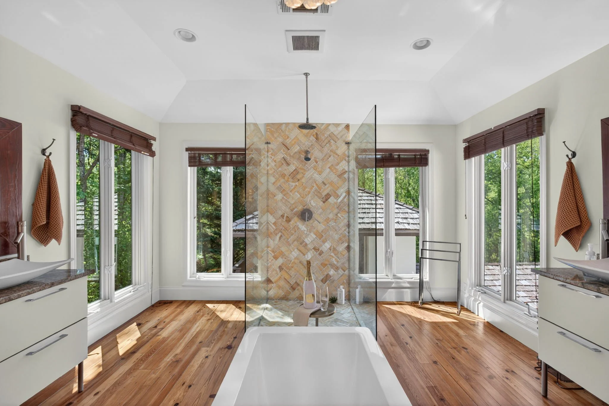 Modern bathroom with large windows, a walk-in shower with a glass partition, a bathtub, wooden floors, and white vanity cabinets.