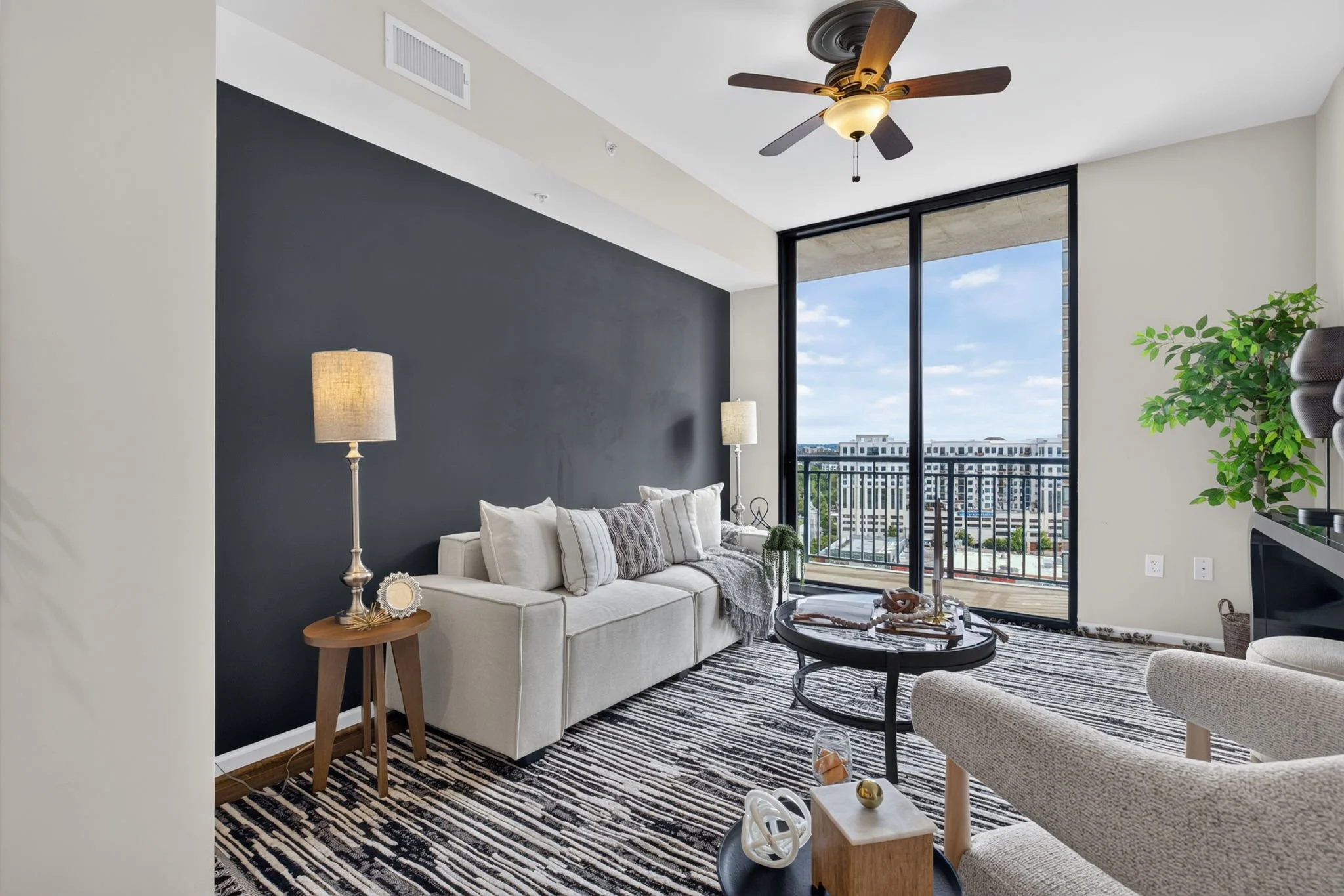 Modern living room with a white sofa, striped rug, black coffee table, boho decor, large window with cityscape view, ceiling fan, and lamps.