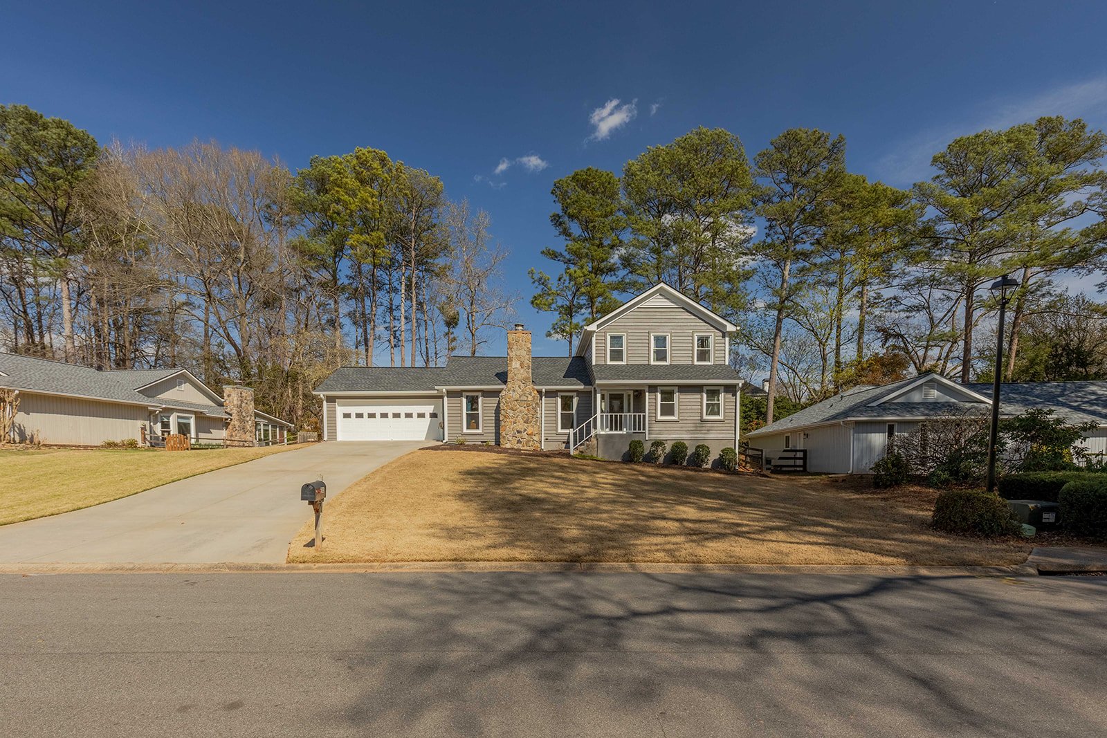 A two-story house with gray siding and a stone chimney, a white garage door, and a front porch with white railing. The house is set on a sloped yard with a concrete driveway, a mailbox, and surrounded by tall trees and neighboring houses. The sky is 