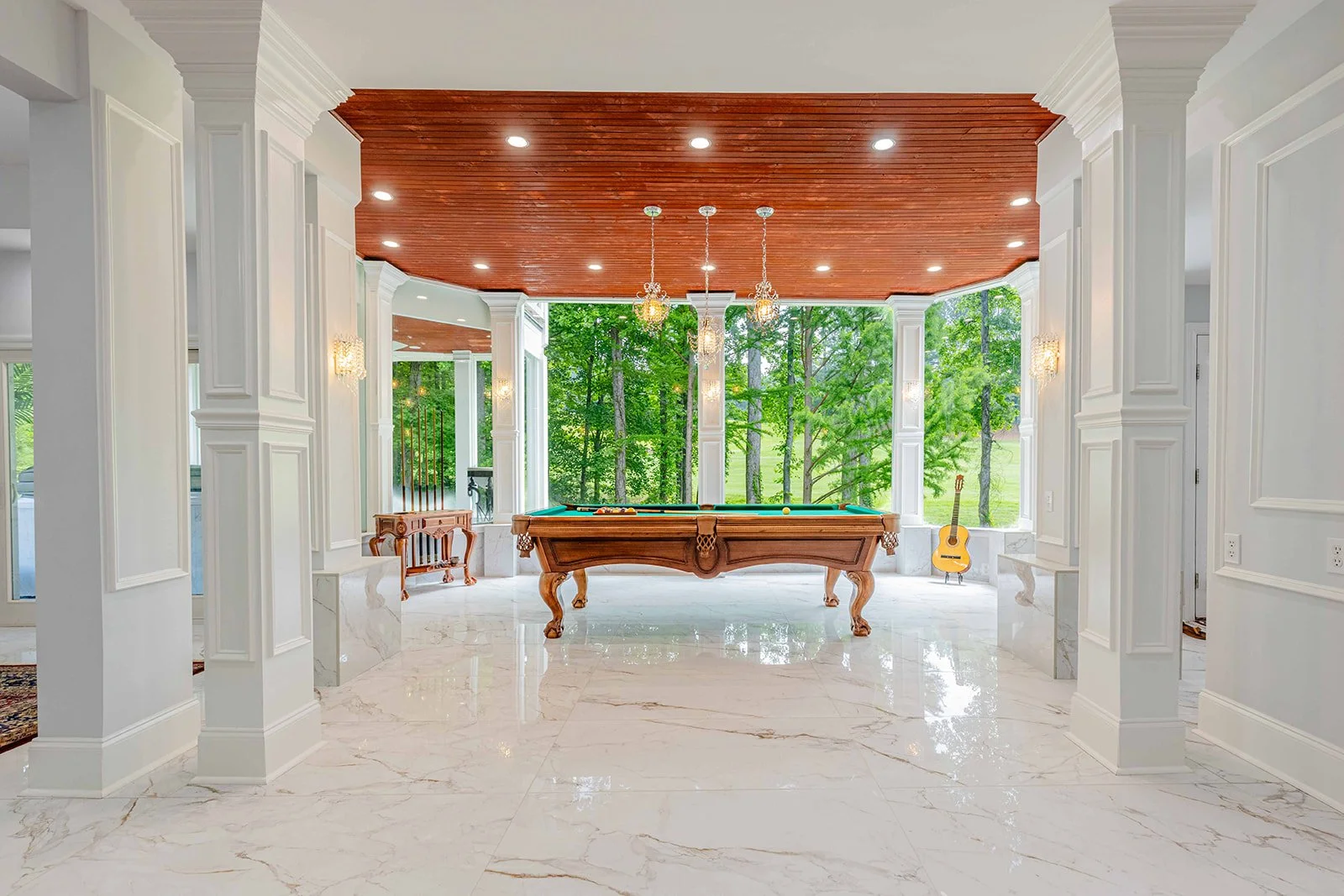 Bright living room with white paneled walls, marble flooring, a wooden ceiling, and large windows showing greenery outside. Features a wooden pool table, a guitar, and chandeliers.